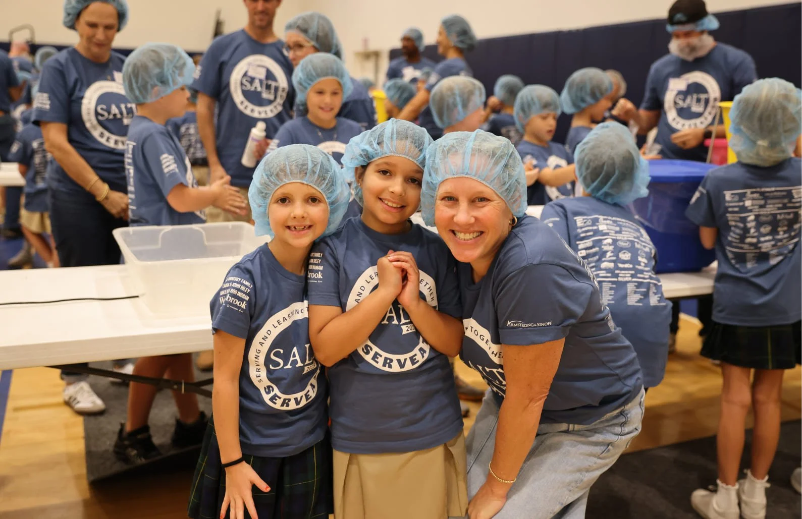 Children and adults wearing blue SALT SERVE shirts and hairnets in a cafeteria or school setting, participating in a community service event.