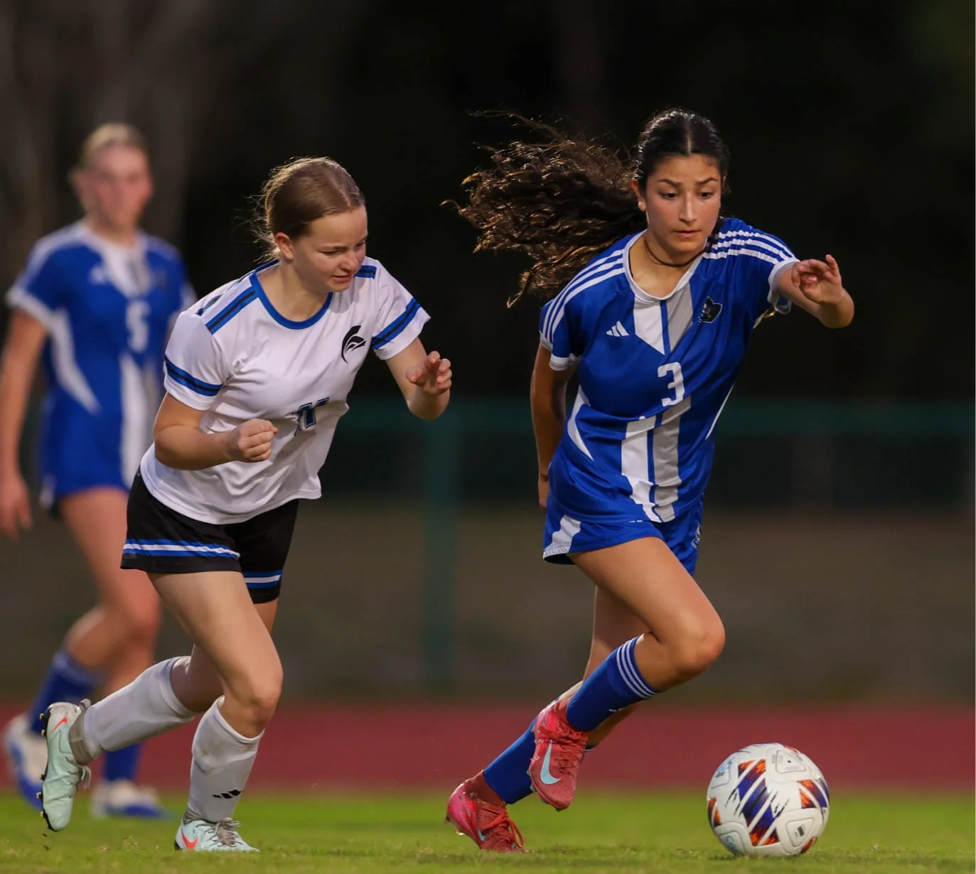 Girls playing soccer on a field, with one player in blue jersey number 3 controlling the ball while others chase behind.