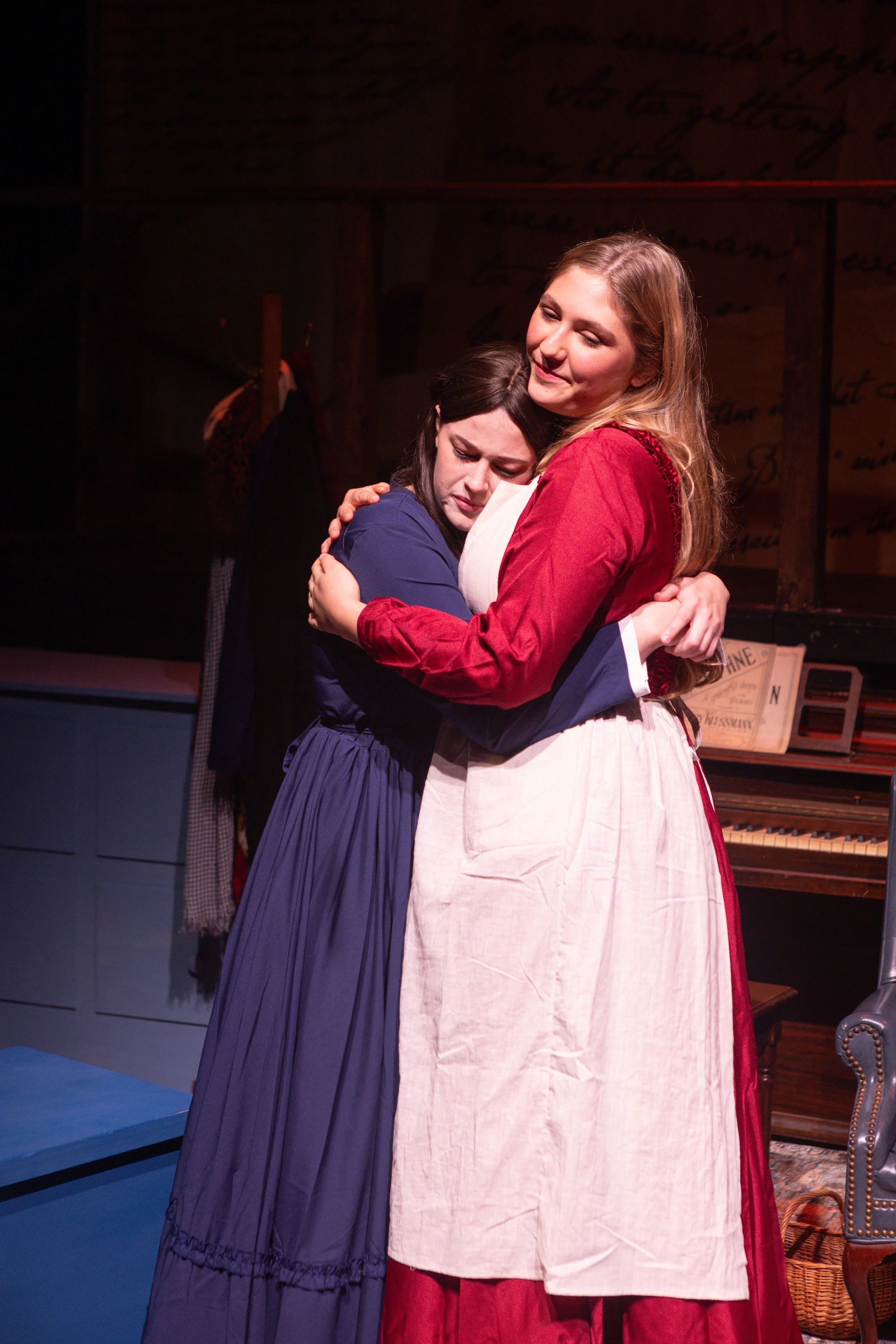 Students embracing in a theatrical performance, with one woman in a blue dress and the other in a red and white dress, on stage with a piano and vintage furniture in the background.