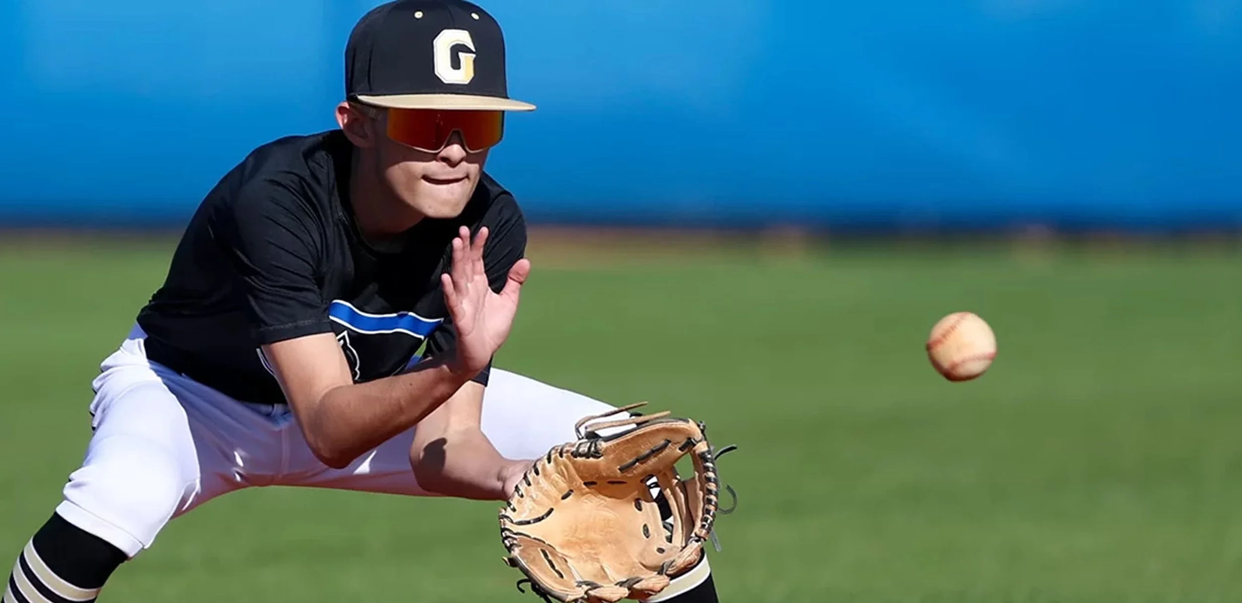 A baseball player in a black jersey, white pants, and a black cap with a letter G, crouching on the field, wearing sunglasses and a baseball glove, with a baseball approaching him in mid-air during daytime.
