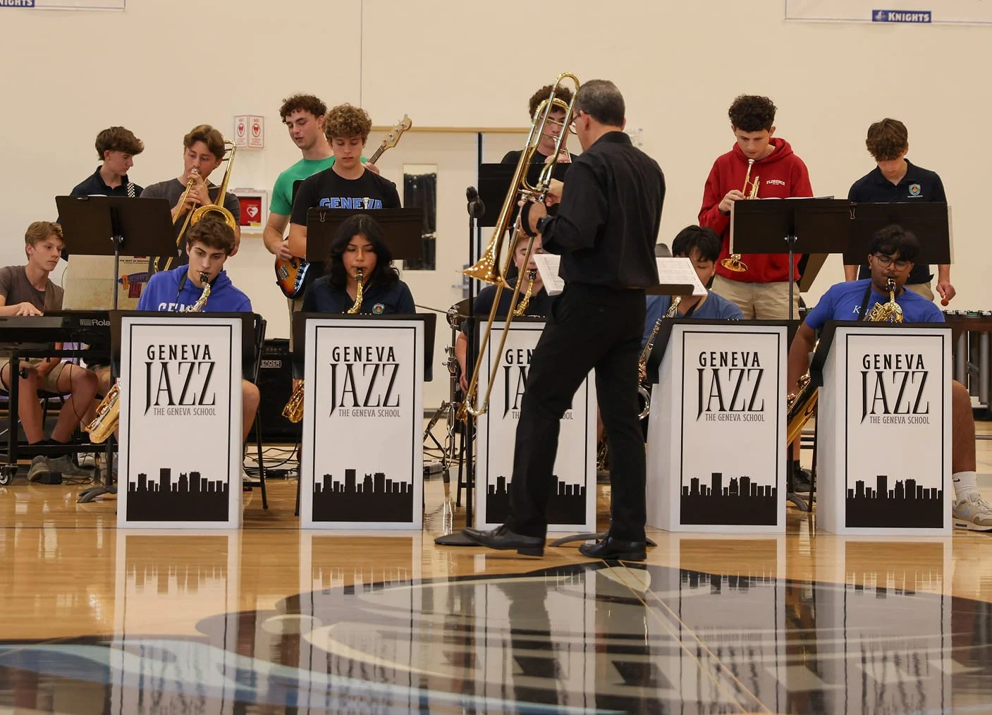 Students in a jazz band performing on stage with a conductor and music stands labeled "Geneva Jazz The Geneva School" at a school event.