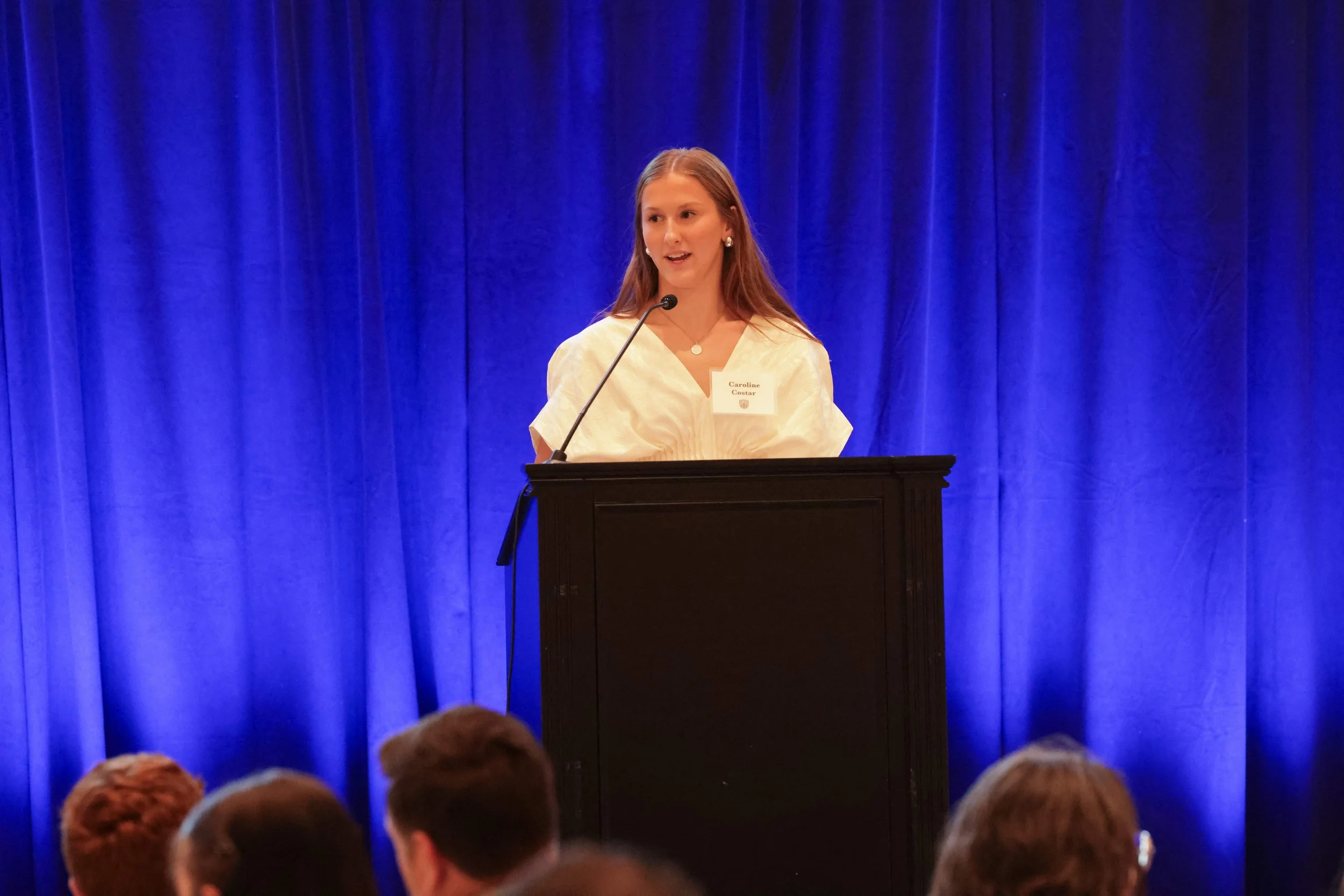 A young woman with long reddish hair speaking at a podium with a microphone in front of a blue curtain backdrop during an event. She wears earrings, a light-colored dress, and has a name tag that reads 'Caroline'.