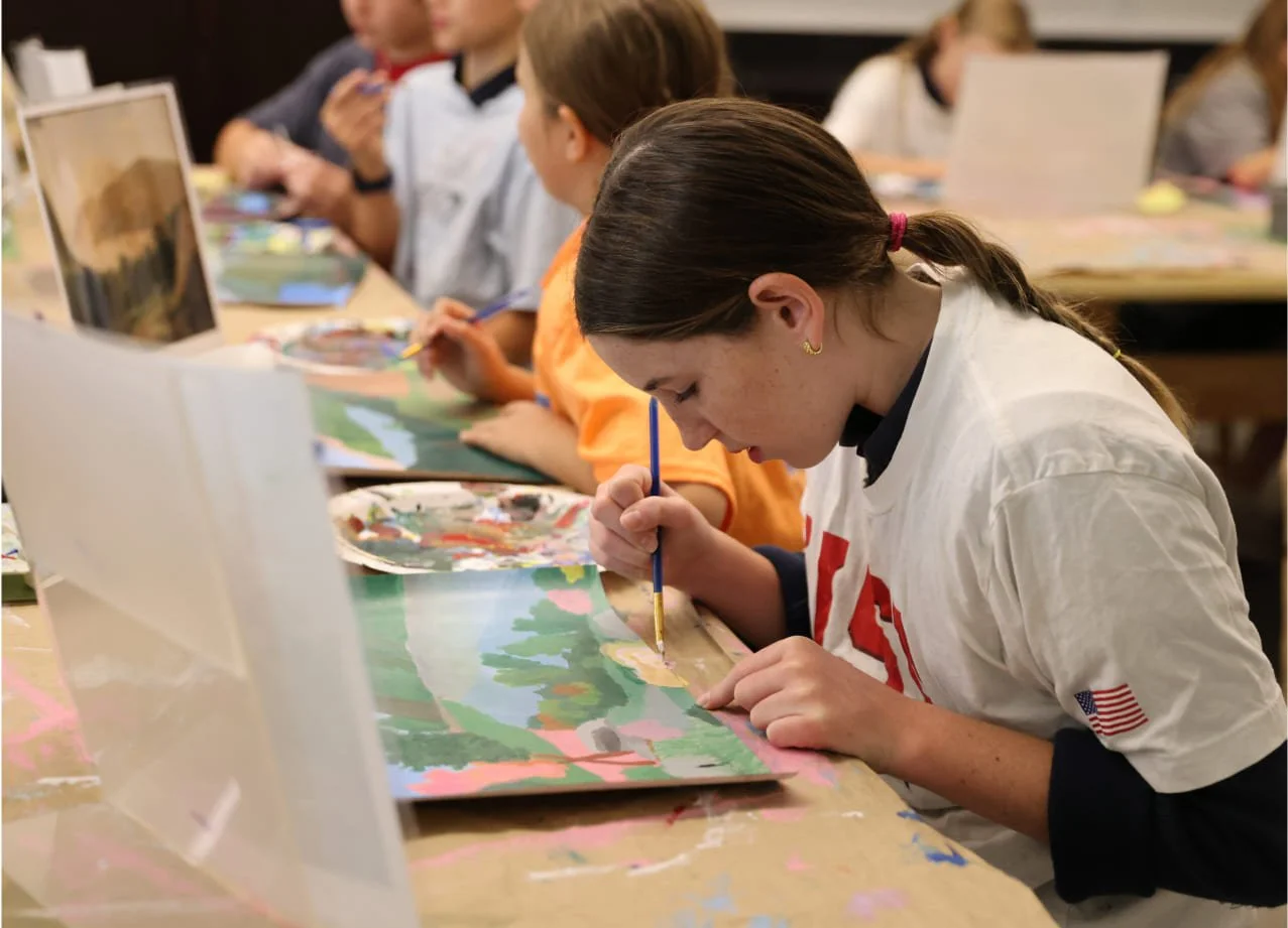 Young girl painting a landscape on paper at an art class, with other children painting nearby.