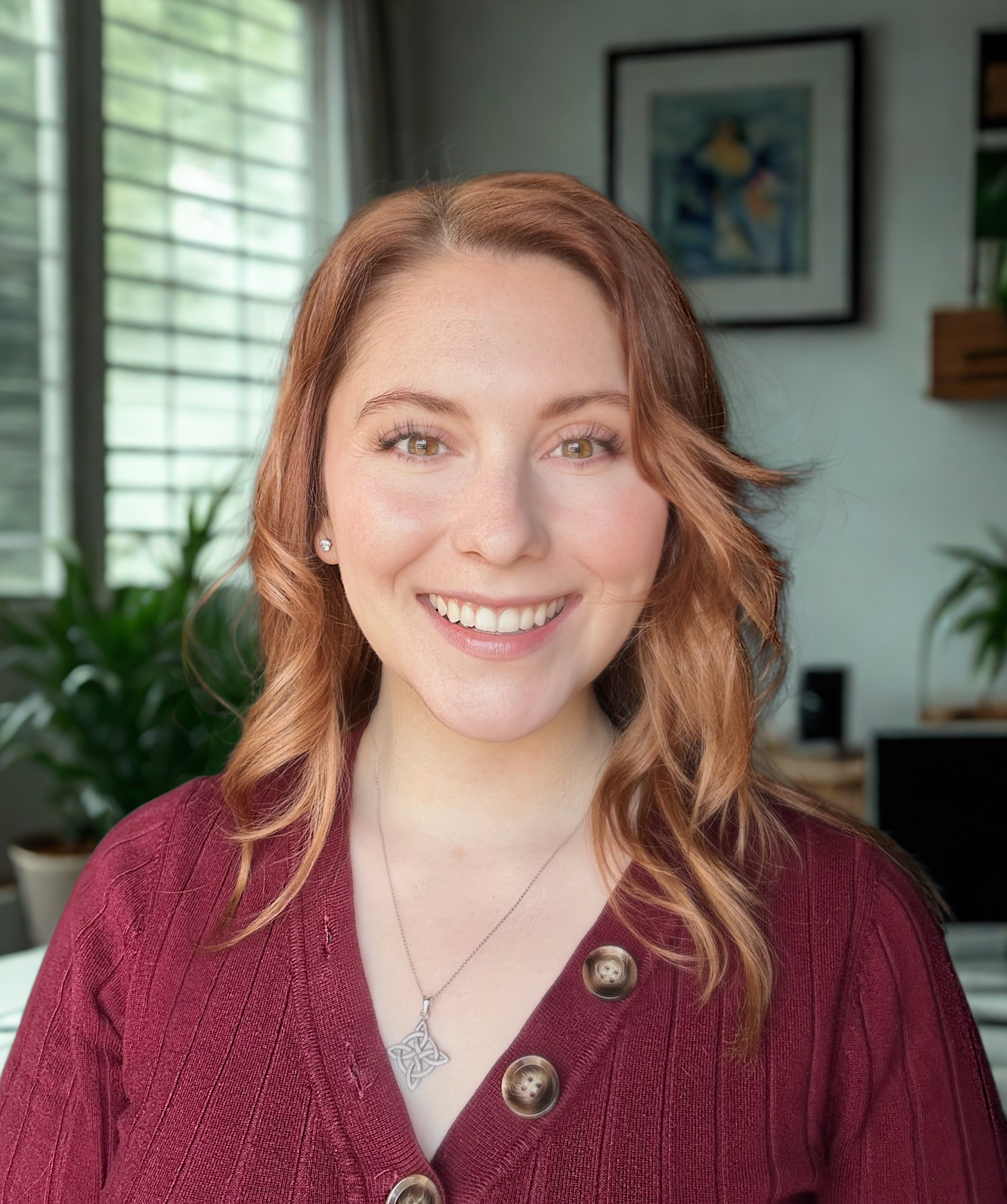 Smiling woman with red hair wearing a maroon sweater with large buttons, a silver necklace, and pearl earrings, in a room with green plants and framed artwork on the wall.