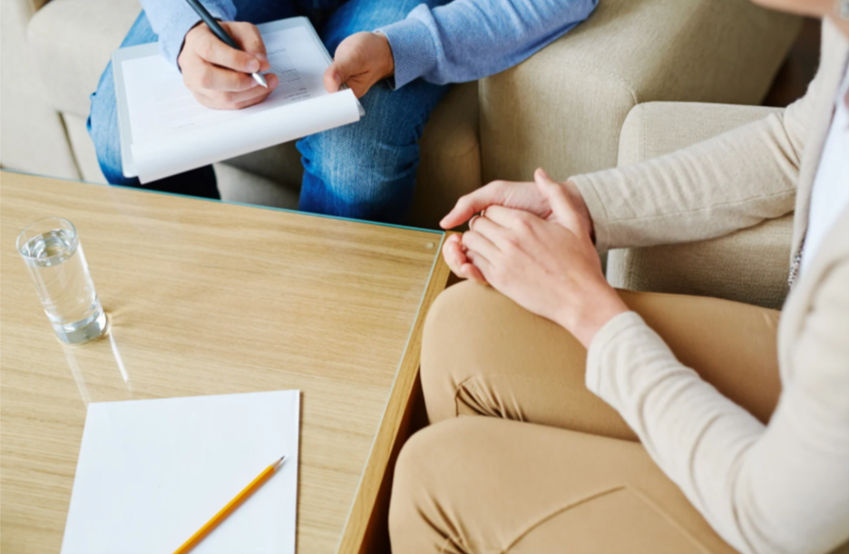 A person sitting on a beige couch during a therapy session, with a therapist taking notes. There is a glass of water and a notepad with a pencil on the wooden table in front.