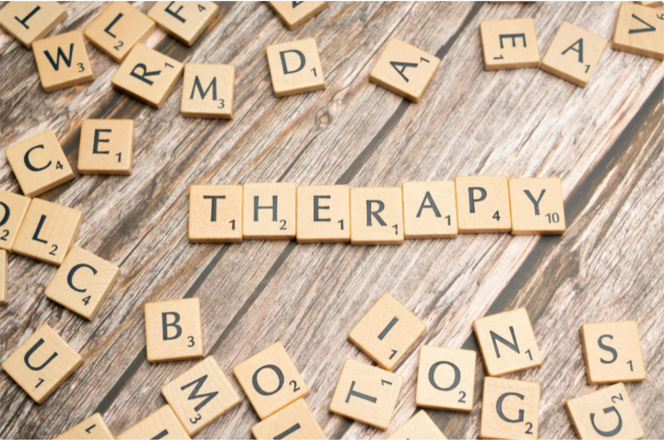 Wooden Scrabble tiles spelling "THERAPY" on a wooden surface surrounded by scattered tiles.
