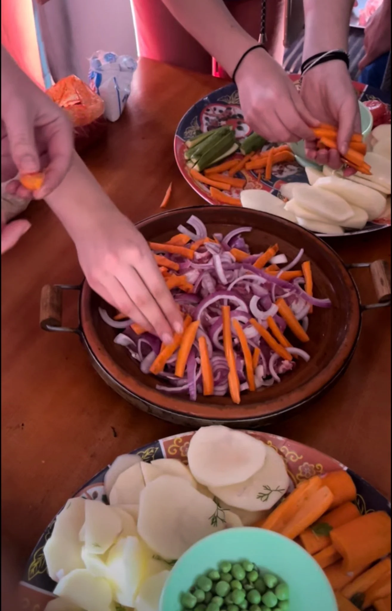 People preparing a vegetable platter with sliced onions, carrots, potatoes, zucchini, and green peas on a wooden table.