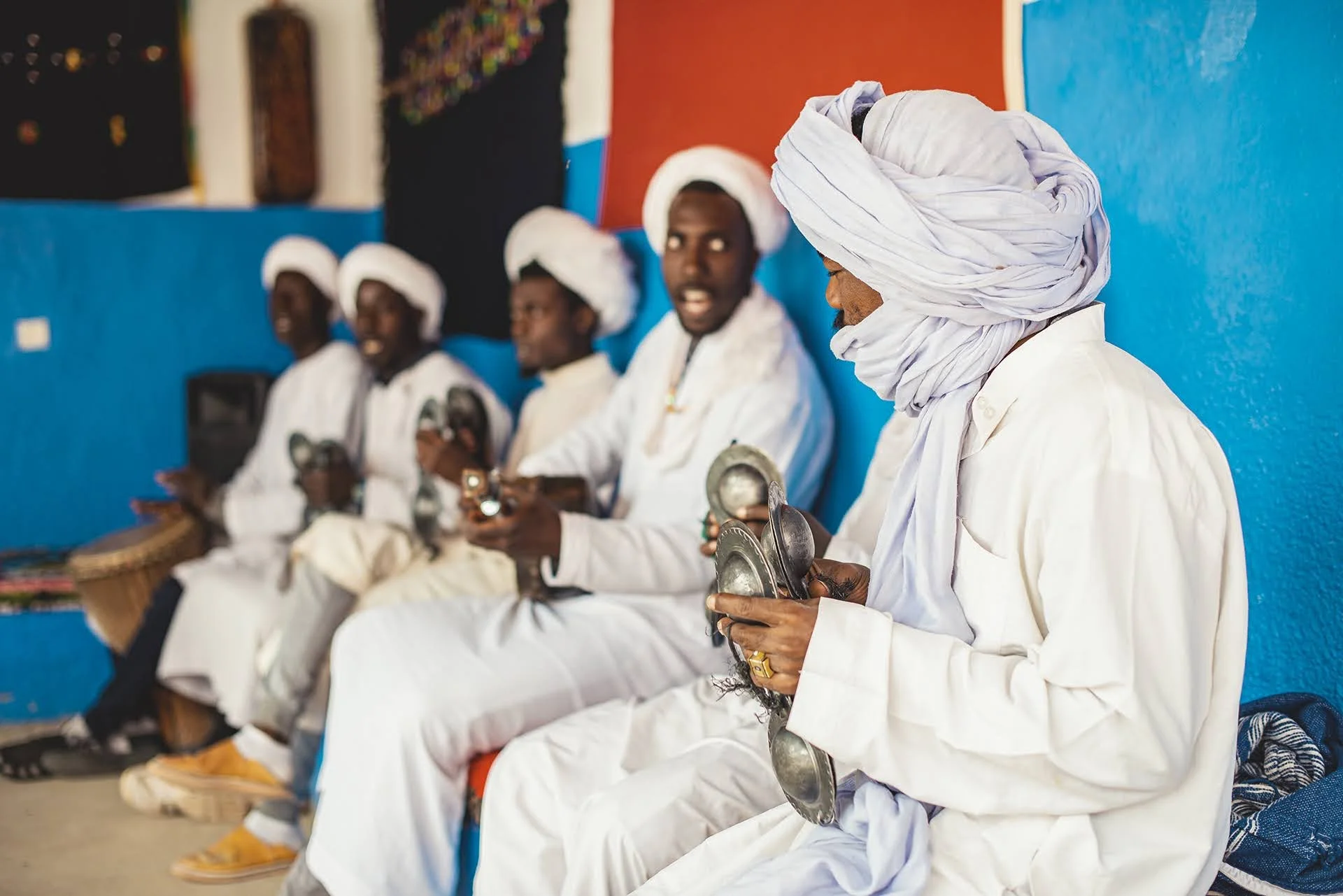 Group of men dressed in traditional white clothing and turbans, sitting in a row against a blue and red wall, holding metal percussion instruments, in a cultural or musical setting.