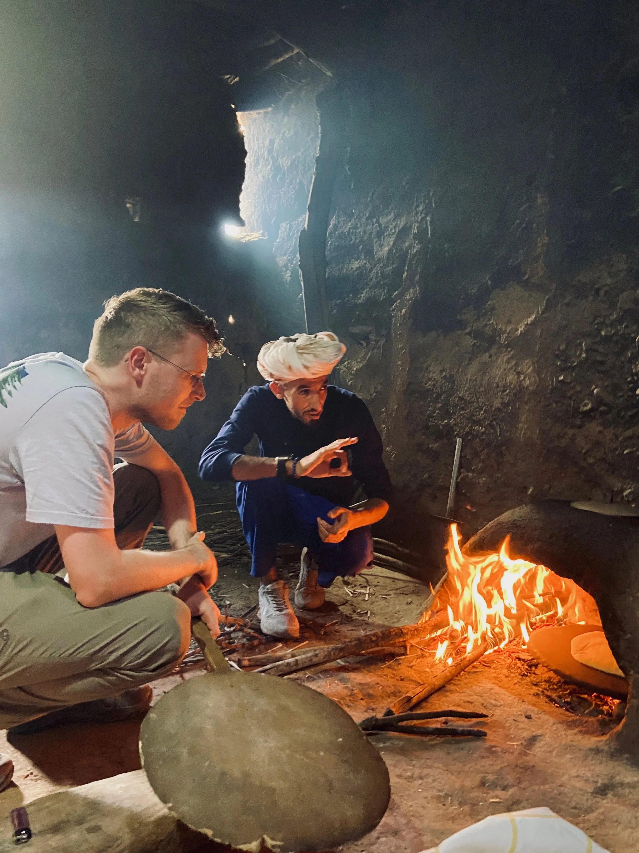 Two men crouching near a small fire inside a dark cave, with one man wearing a turban and making a gesture with his hand.