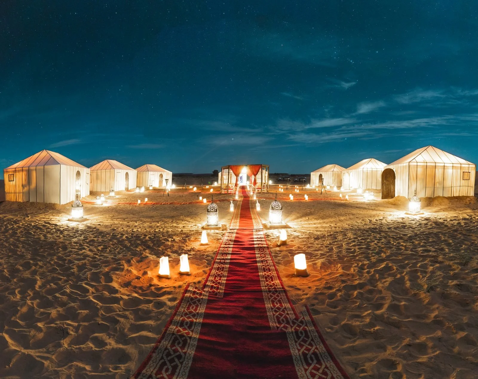 Candle-lit desert camp at night with white tented rooms, a red carpet walkway, lanterns on the sand, and a star-filled sky overhead.t, mountains in the background.
