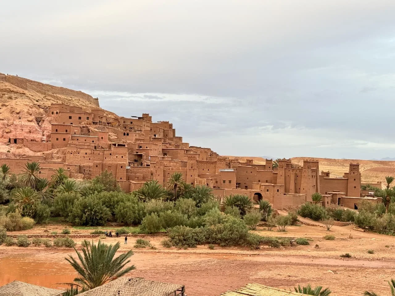 Ancient ksar of Aït Benhaddou in Morocco, with adobe buildings rising above palm trees and the surrounding desert landscape.