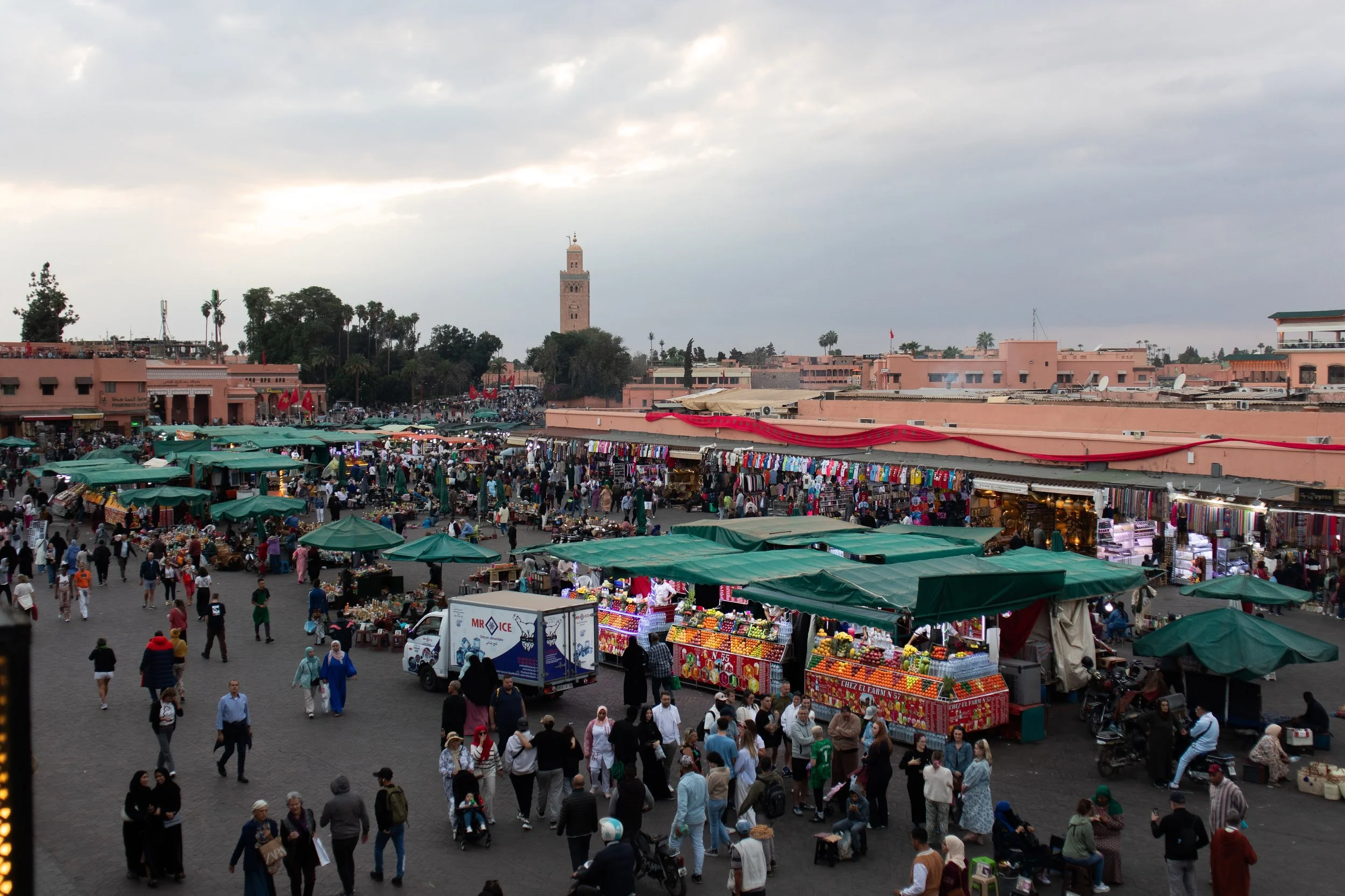 A lively street market with numerous stalls covered by green canopies, crowded with shoppers and vendors. The market is set in a city with pink-colored buildings, a tall minaret tower in the background, and a cloudy sky overhead.