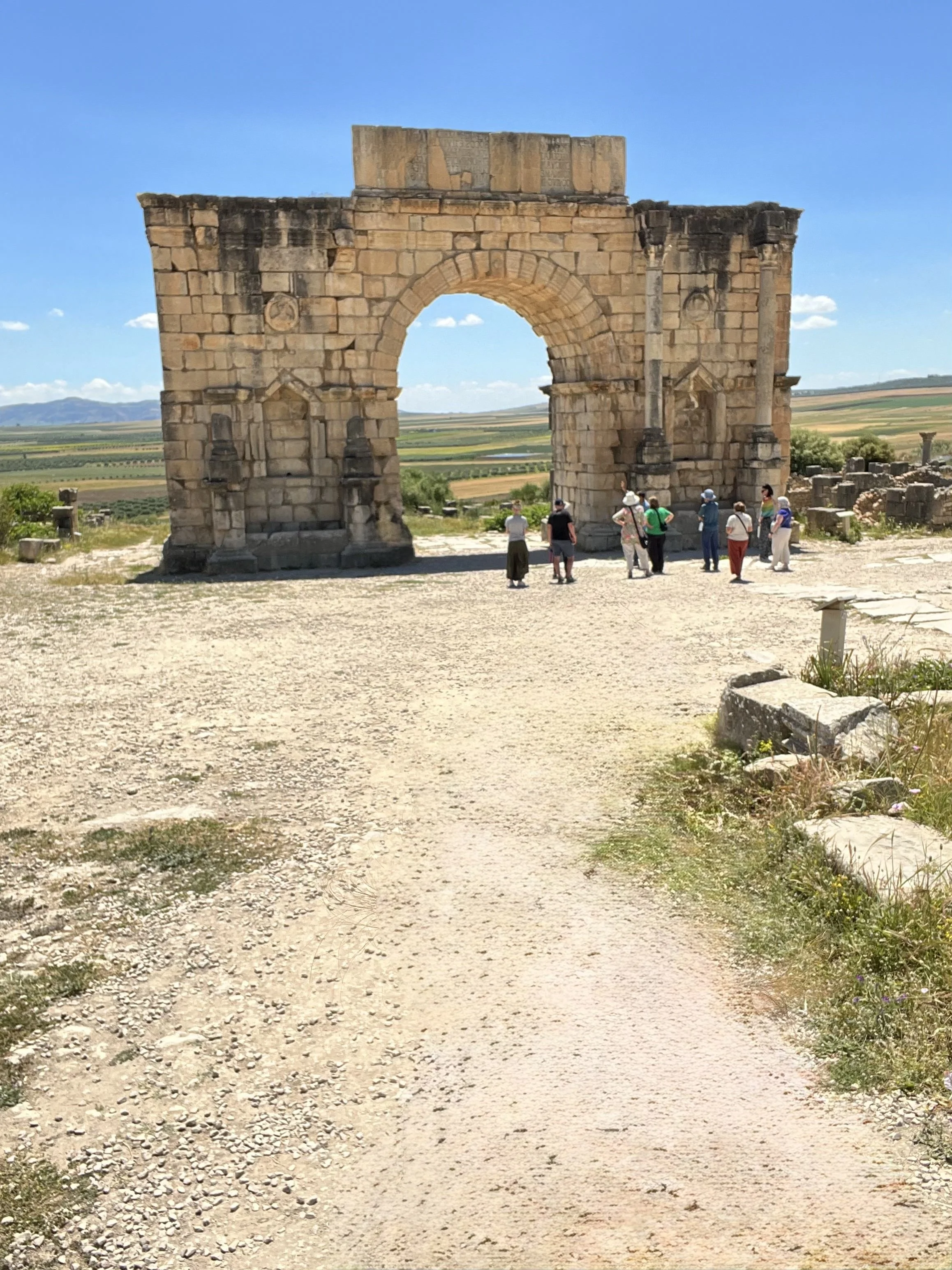 Ancient stone arch ruins with tourists nearby, set against a bright blue sky and rural landscape.
