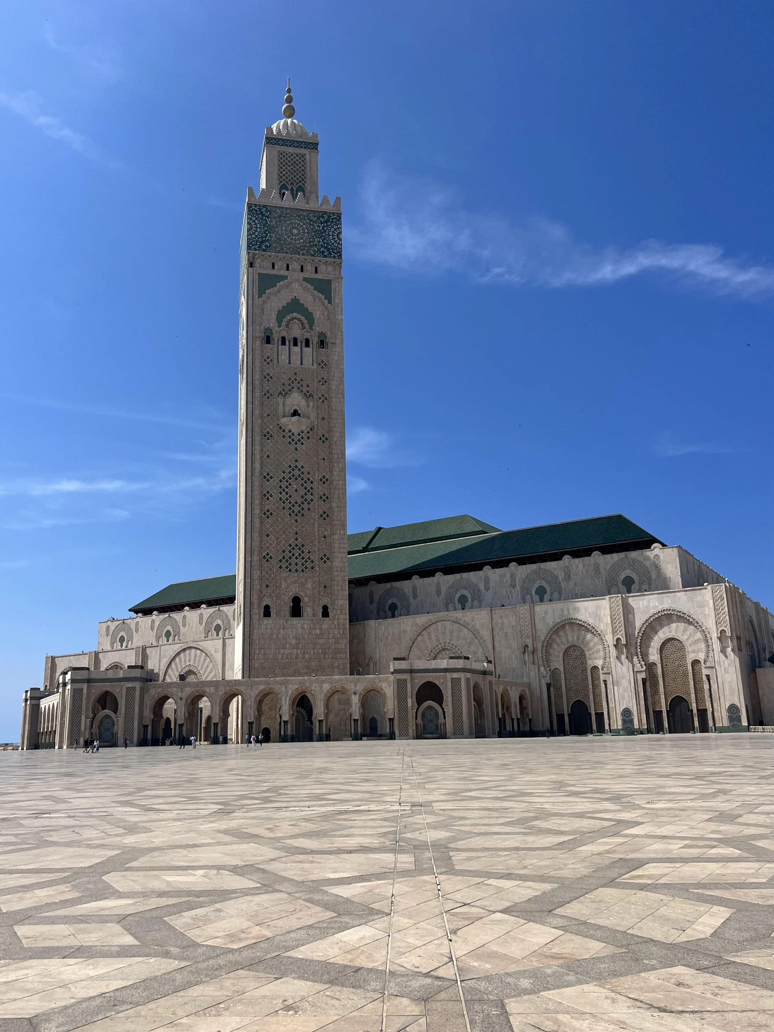 A mosque with a tall minaret and green roof, standing on a large open courtyard with patterned tile flooring, under a clear blue sky.