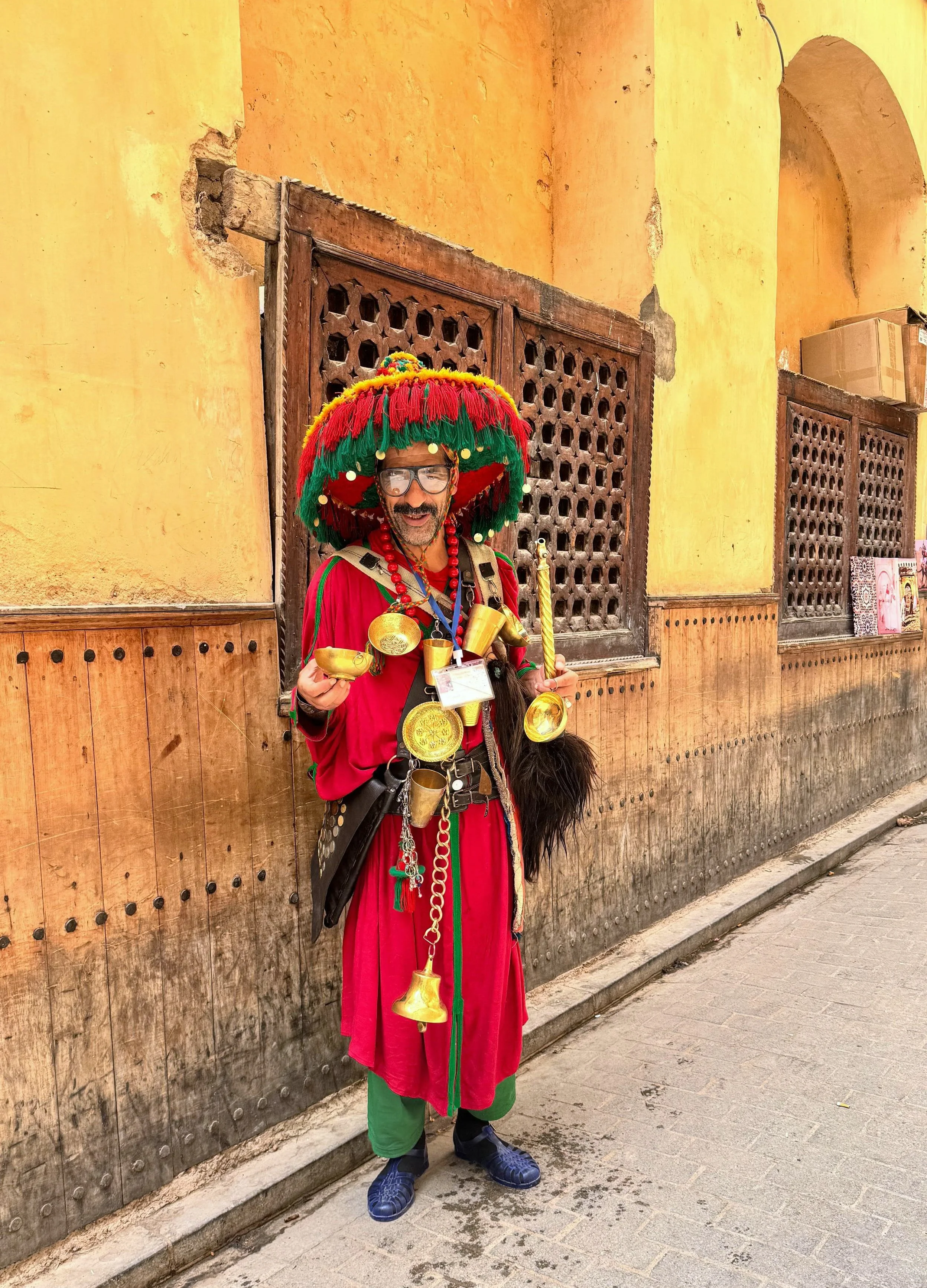 A man dressed in colorful, traditional Mexican attire, including a large red and green hat decorated with pom-poms, gold medals and bells, standing against a yellow stucco wall with wooden lattice windows.