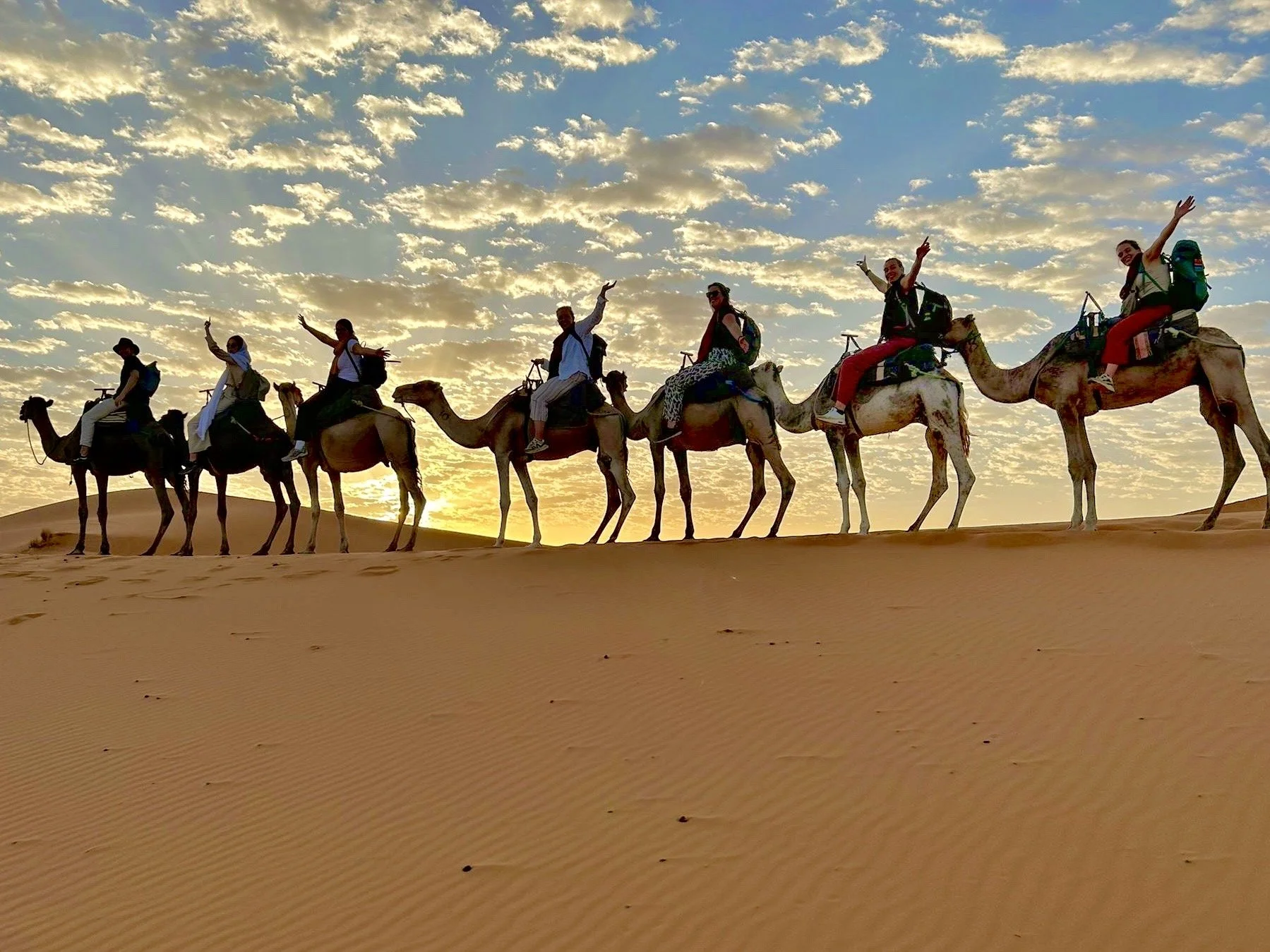 A group of people riding camels in a desert at sunset, all raising one arm and smiling.