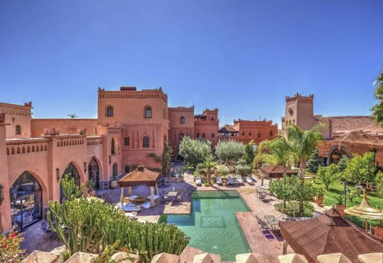 A luxurious hotel with a pinkish stone exterior, surrounded by lush trees, a swimming pool, and outdoor lounging areas, under a clear blue sky.