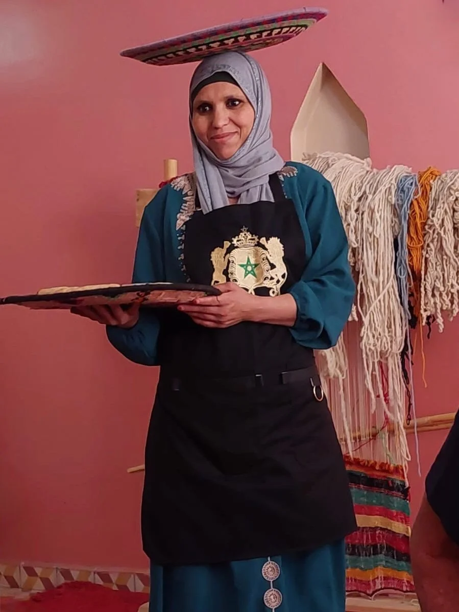 A woman wearing a hijab with a cloth on her head and an apron with a crest, balancing a colorful tray on her head, standing in front of a pink wall with woven textiles hanging in the background.