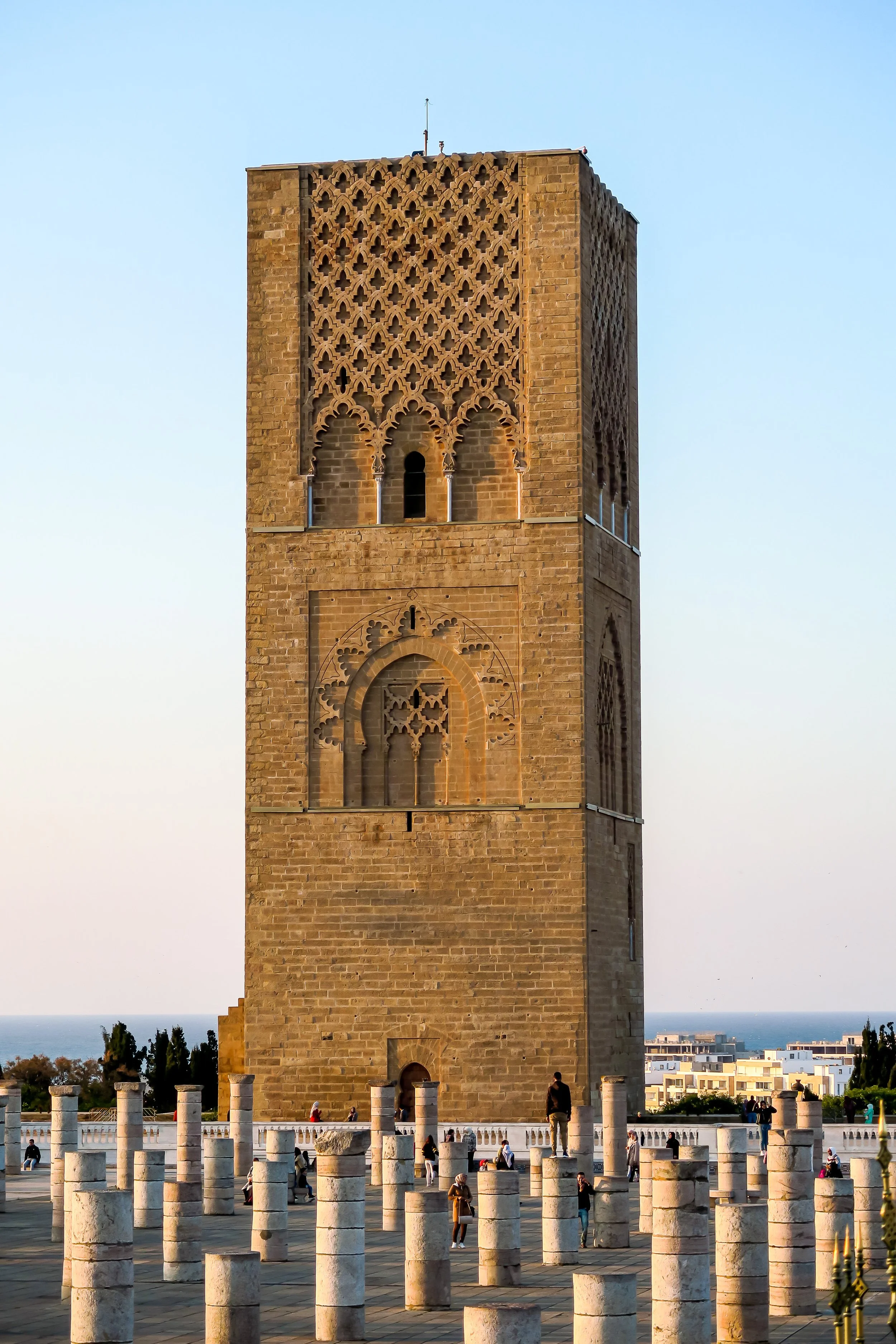 A tall historic stone tower with intricate lattice and arched windows, surrounded by stone pillars, people, and a cityscape background.