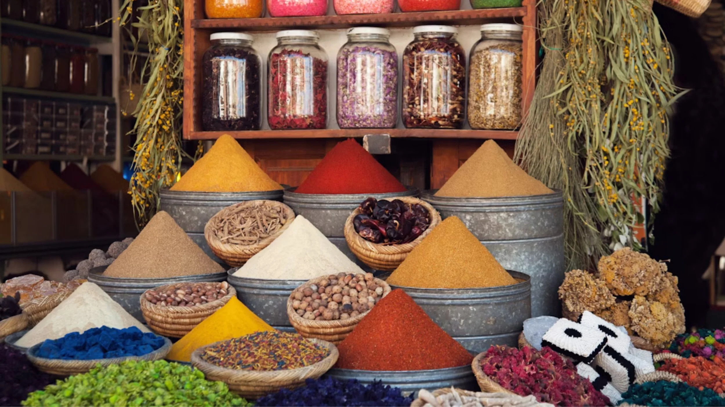 Display of colorful spices in metal containers, hanging herbs, and jars of dried items on a market stall.