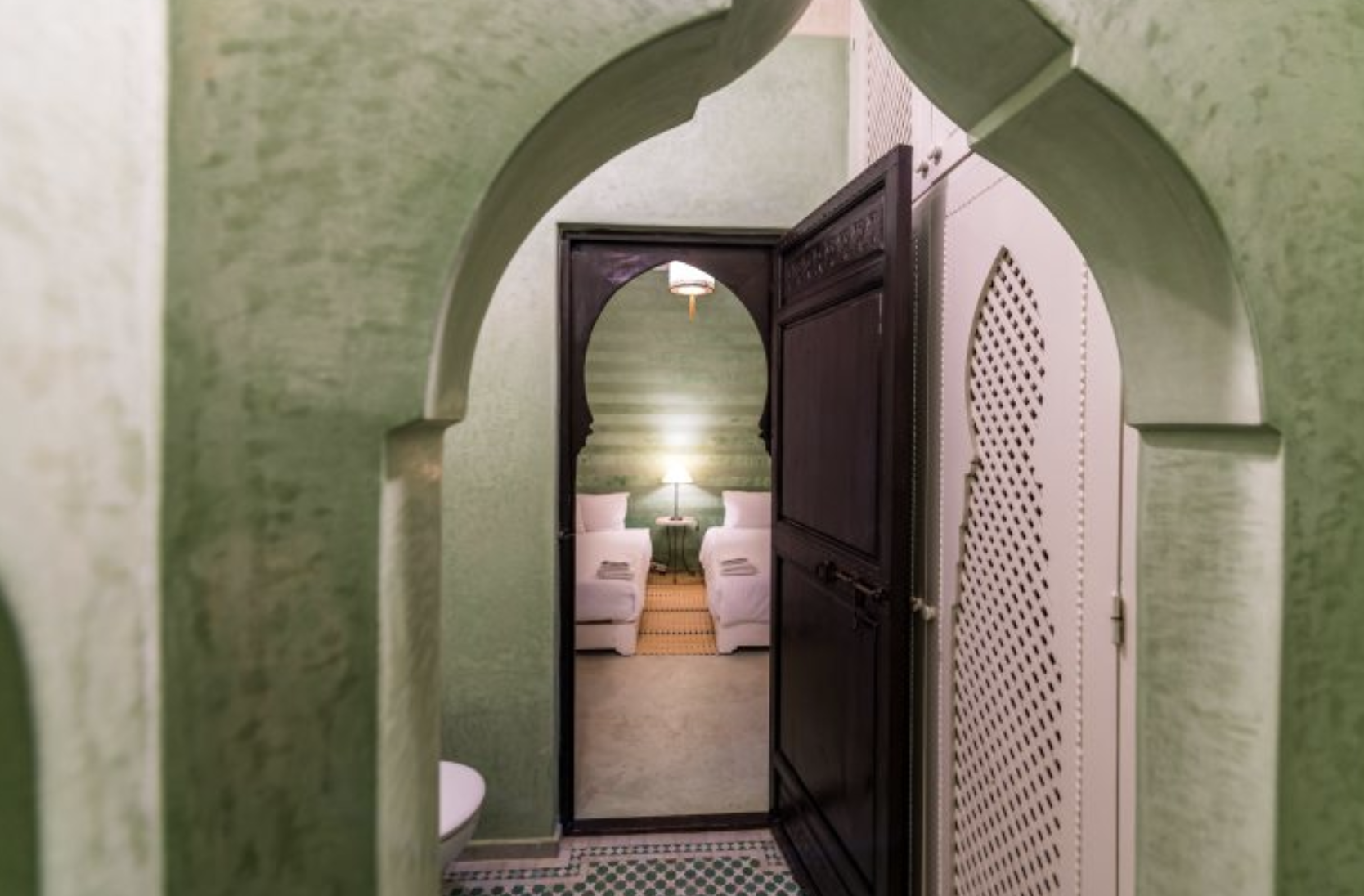 View through an arched green hallway into a room with two white couches facing each other, a small lamp on a side table, and a striped wall behind.