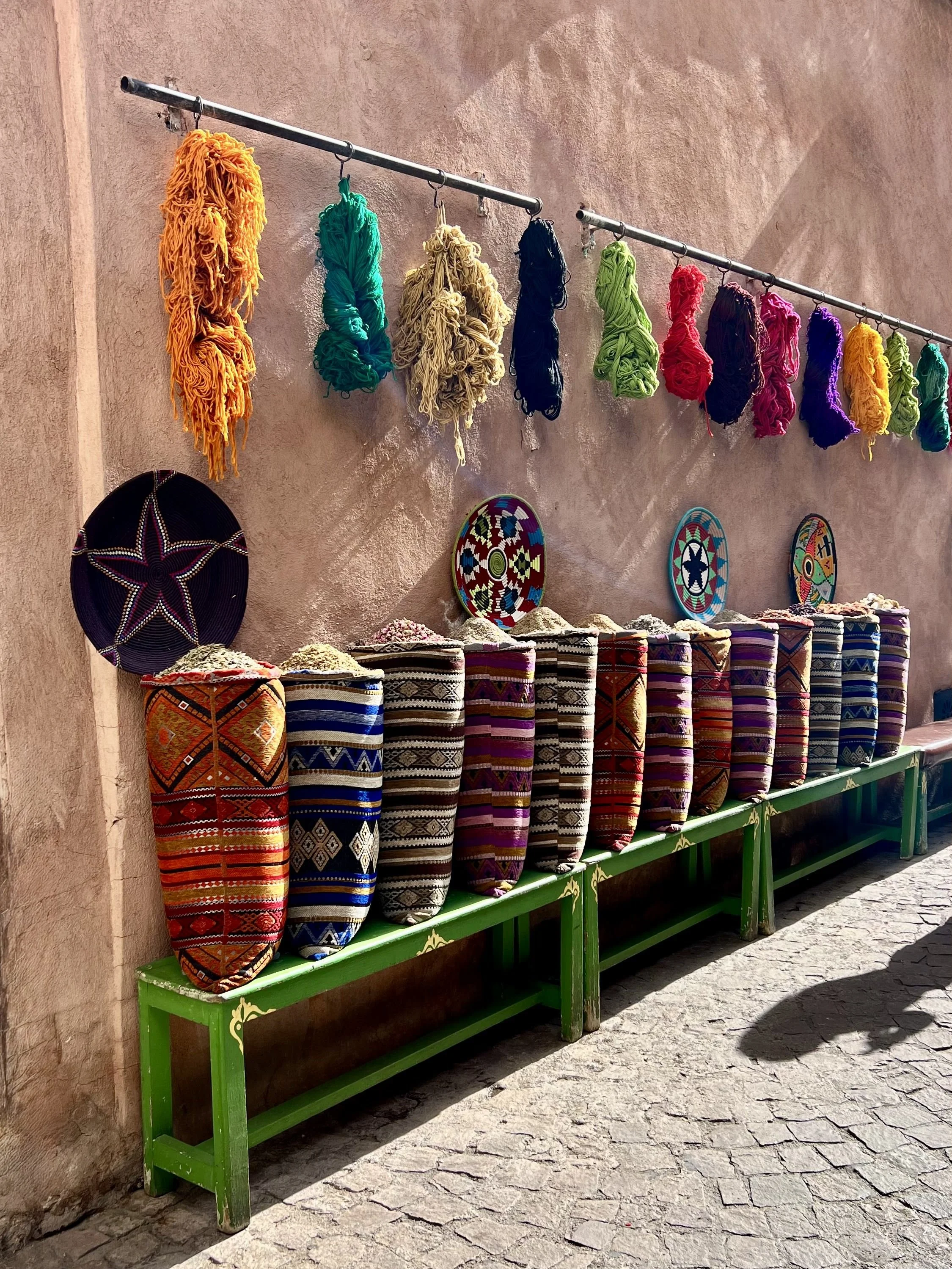 Colorful yarn balls hanging from a metal rod, decorated woven baskets on a green wooden bench, and painted decorative plates on a pink textured wall.