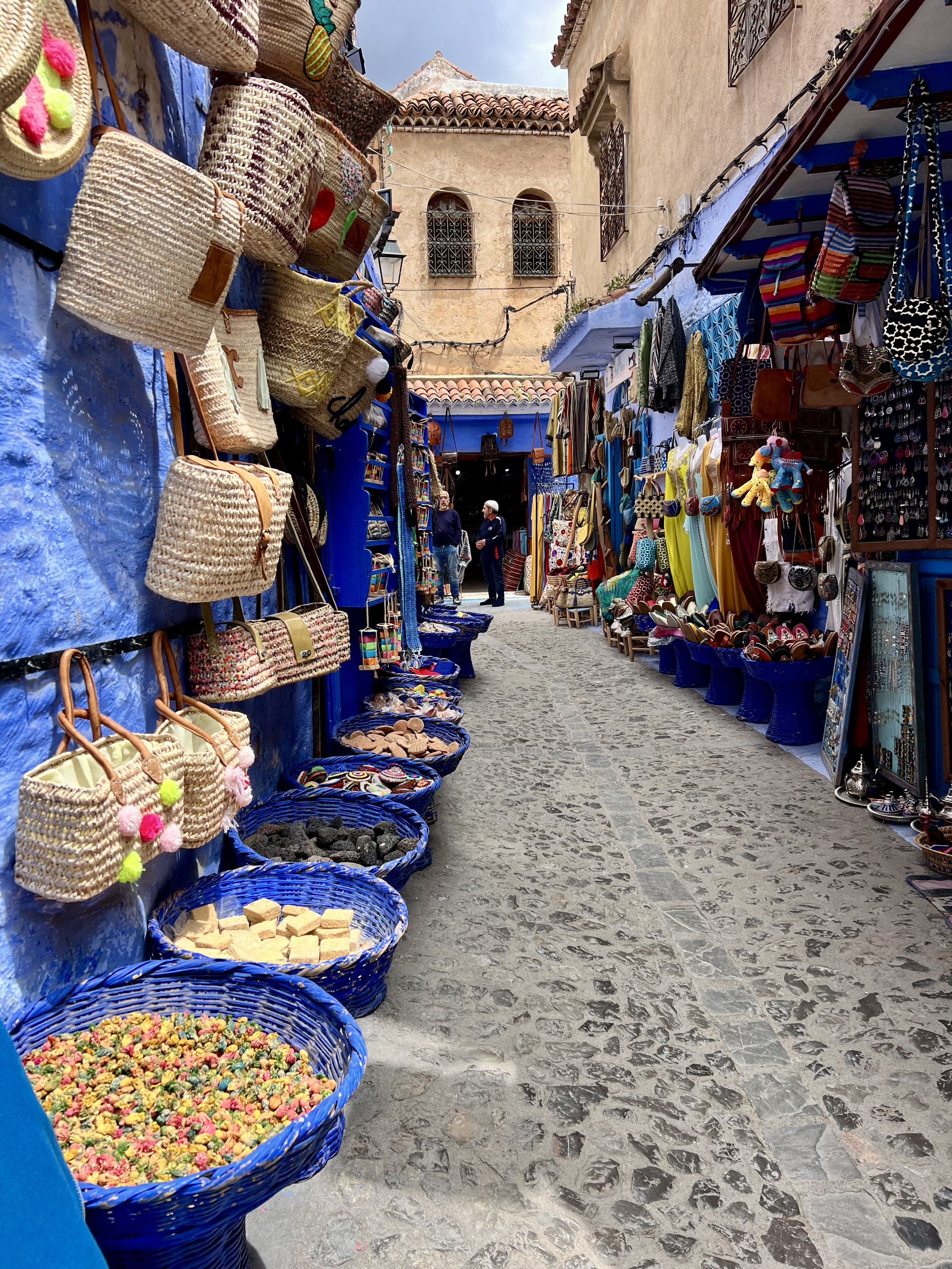 A narrow street lined with vendor stalls selling colorful bags, jewelry, and souvenirs, with a cobblestone sidewalk and rustic buildings in the background.