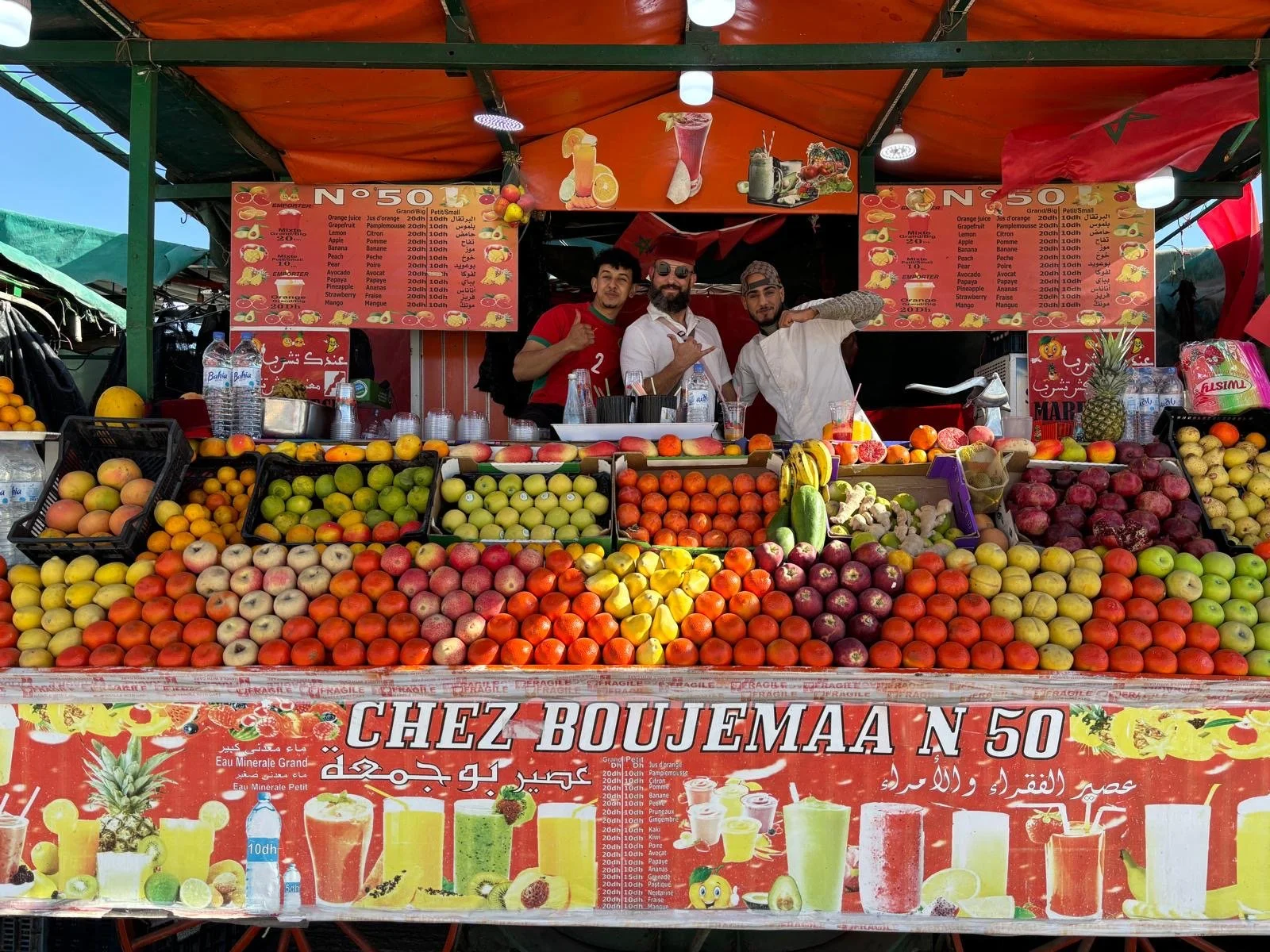A vibrant outdoor fruit stand with various fresh fruits including apples, oranges, bananas, pineapples, and melons on display. Three men are standing behind the stand, smiling and posing for the camera. The stand has red and orange signage with a men