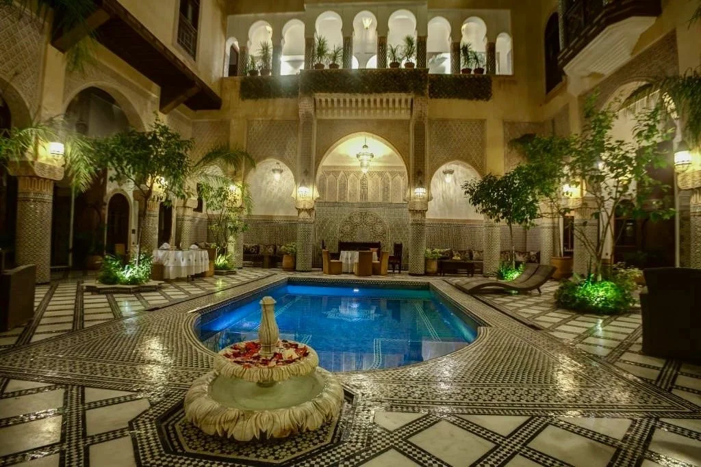 Indoor courtyard with a swimming pool, ornate fountain with petals, lush plants, and elegant seating areas.