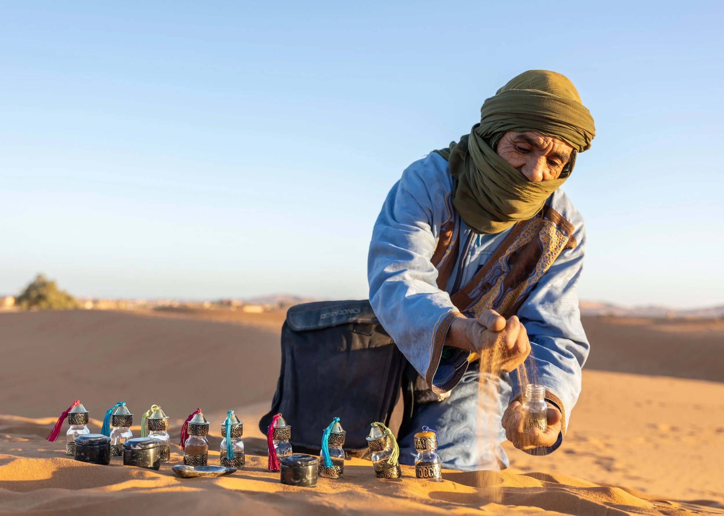 A man wearing traditional desert clothing, including a scarf covering his face and head, arranging small decorative lamps on sand dunes during sunset in a desert landscape.