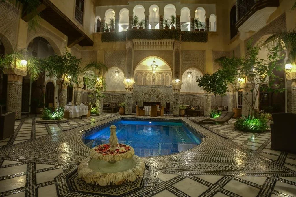 Interior courtyard of a traditional Moroccan riad with a central small swimming pool, decorative mosaic tile flooring, lush green trees, and ornate arches and lanterns.