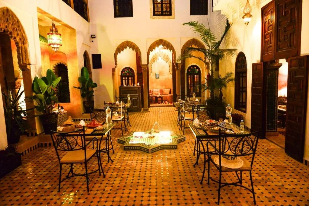 Warm courtyard at Riad Almazhar in Rabat, with patterned tile floors, small café tables, plants, and soft lantern lighting creating a calm, intimate setting.