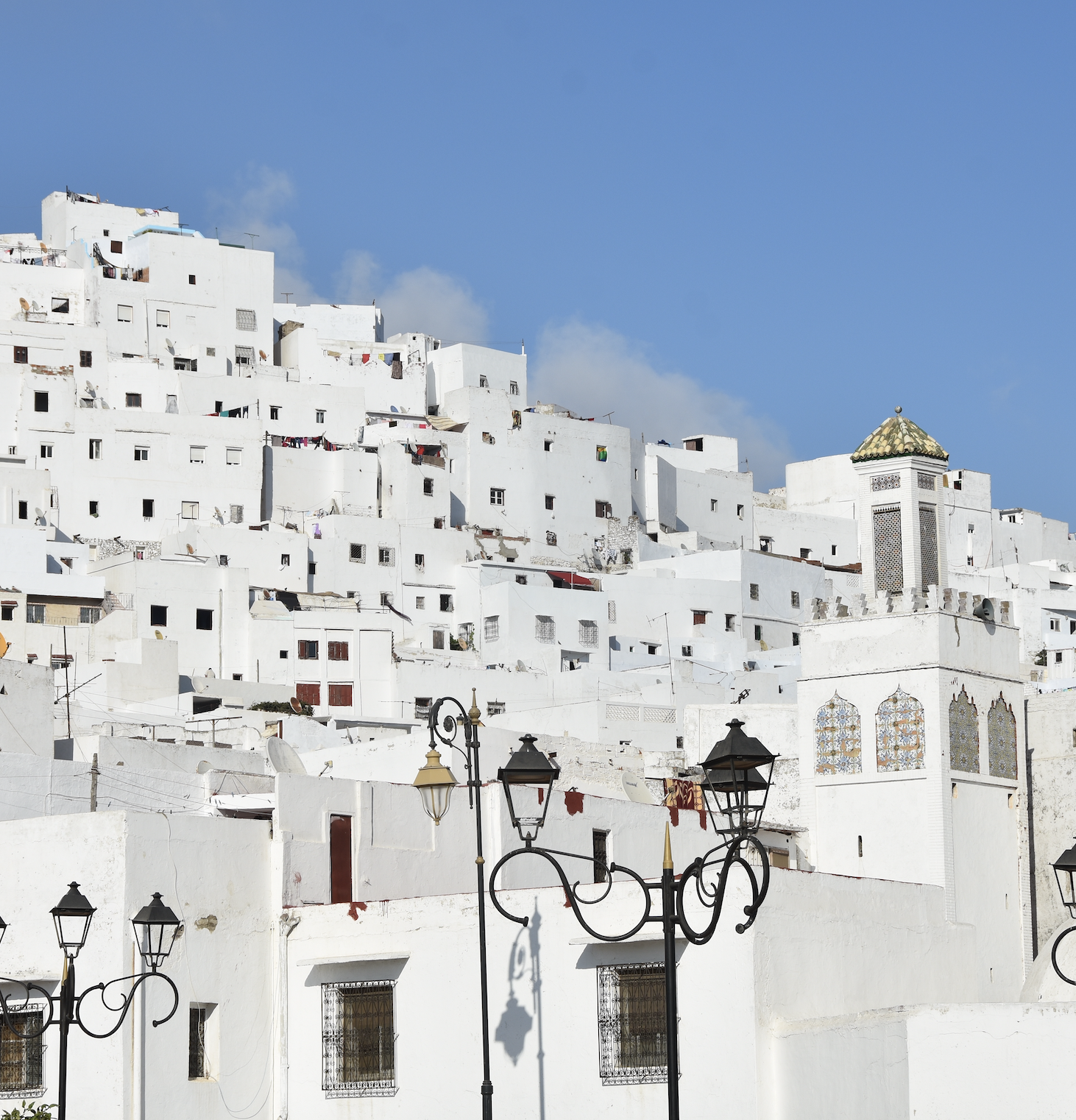A hillside view of white Mediterranean-style buildings with simple geometric shapes, a blue sky, and street lamps in the foreground.
