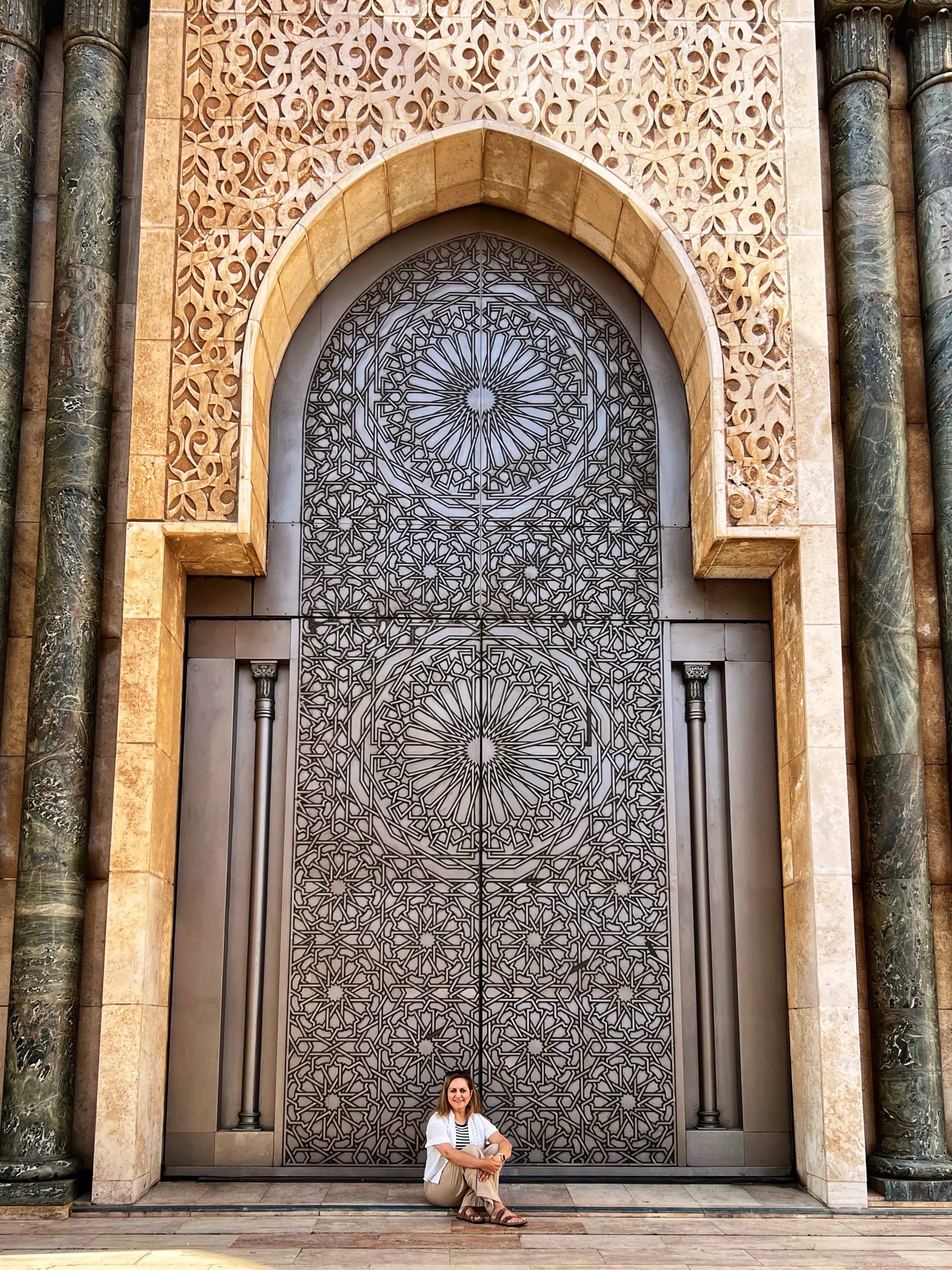A woman sitting on the ground in front of a large, ornate metal door with intricate geometric patterns, surrounded by marble columns and decorative stonework.
