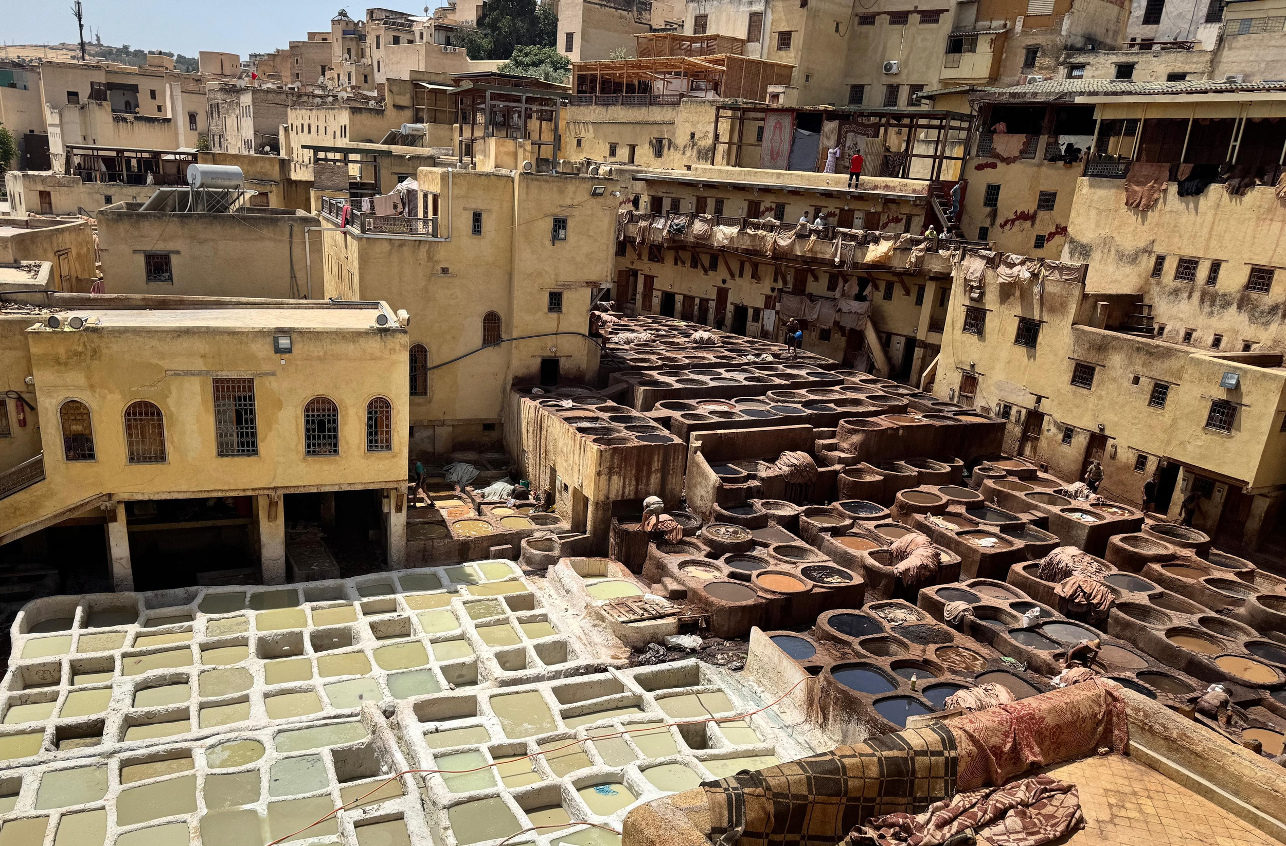 A view of traditional tanneries with large stone and concrete vats filled with various colored liquids used for leather processing, surrounded by old yellowish buildings in an urban area.