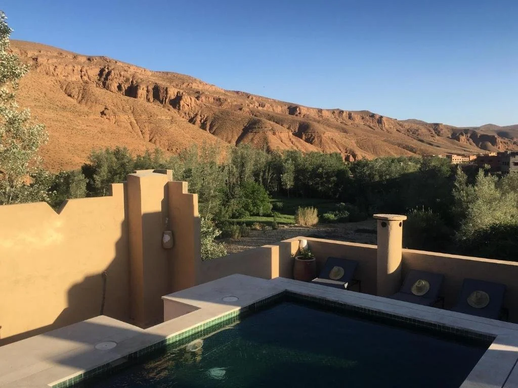 Terrace pool overlooking the Dades Valley, with palm trees, lush greenery, and dramatic red rock cliffs under a clear blue sky.