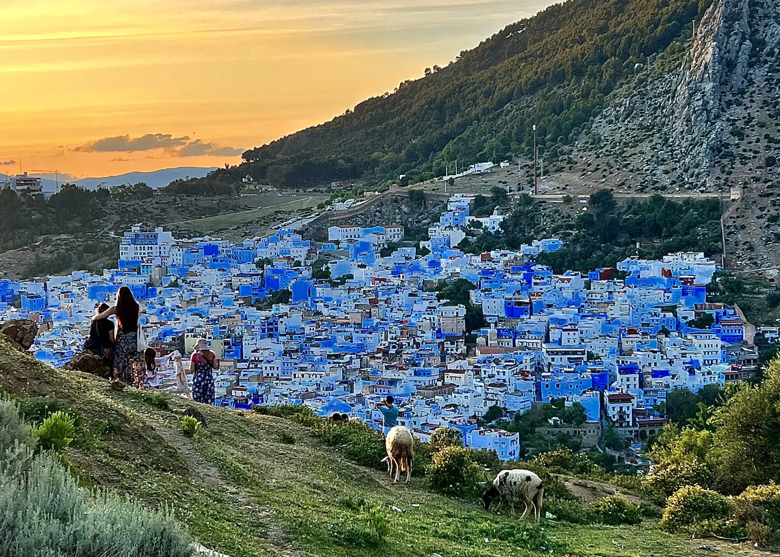 A hillside town with many blue-painted buildings, with a sunset sky and mountain background. In the foreground, a woman with a hat and a backpack stands with a goat, and other women are taking photos of the scenic view.