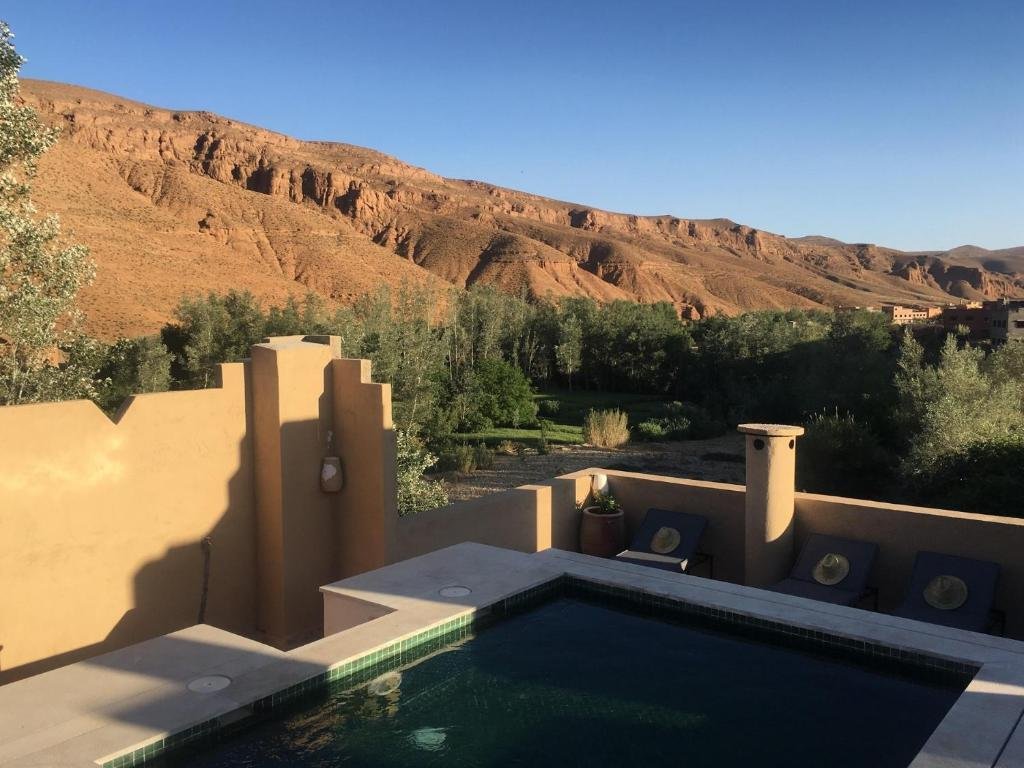Terrace pool overlooking the Dades Valley, with palm trees, lush greenery, and dramatic red rock cliffs under a clear blue sky.
