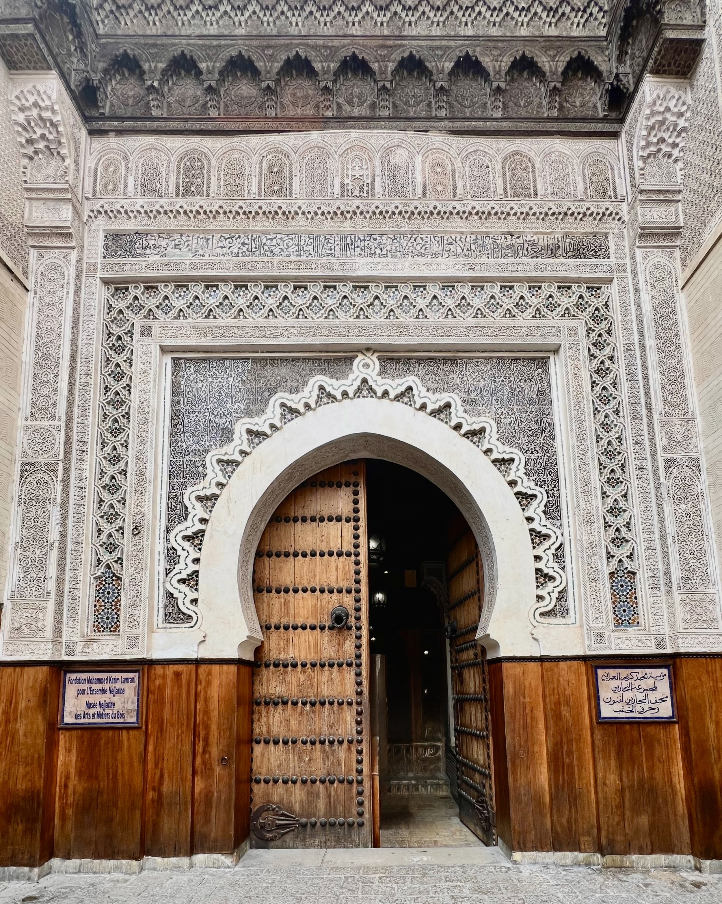 Ornate Moroccan entrance with carved wood, stonework, Arabic inscriptions, and traditional architectural details.