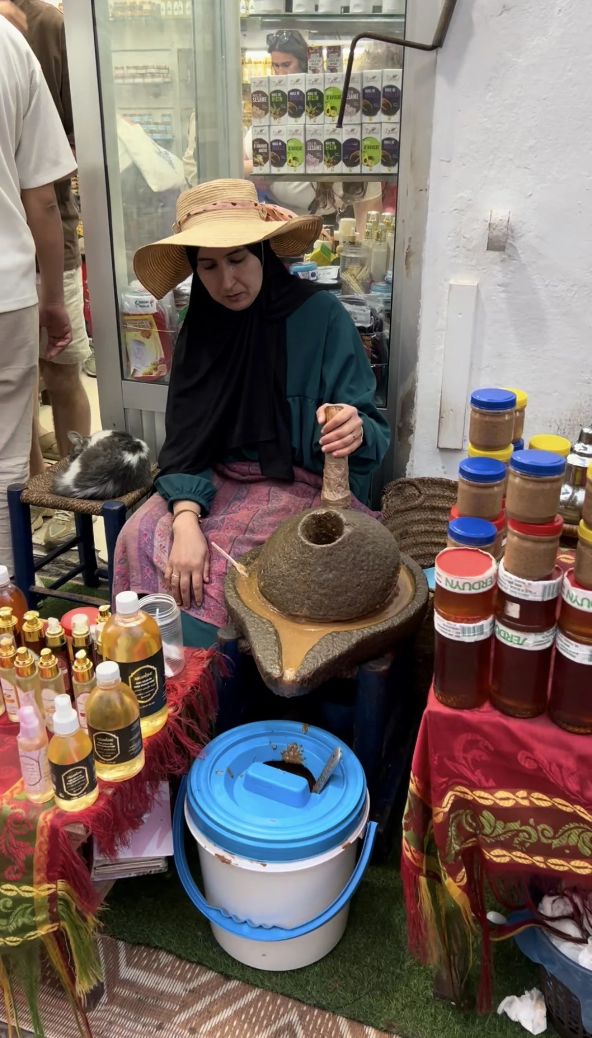 Woman making argan oil by hand in Morocco using traditional grinding methods, surrounded by natural products and market goods.