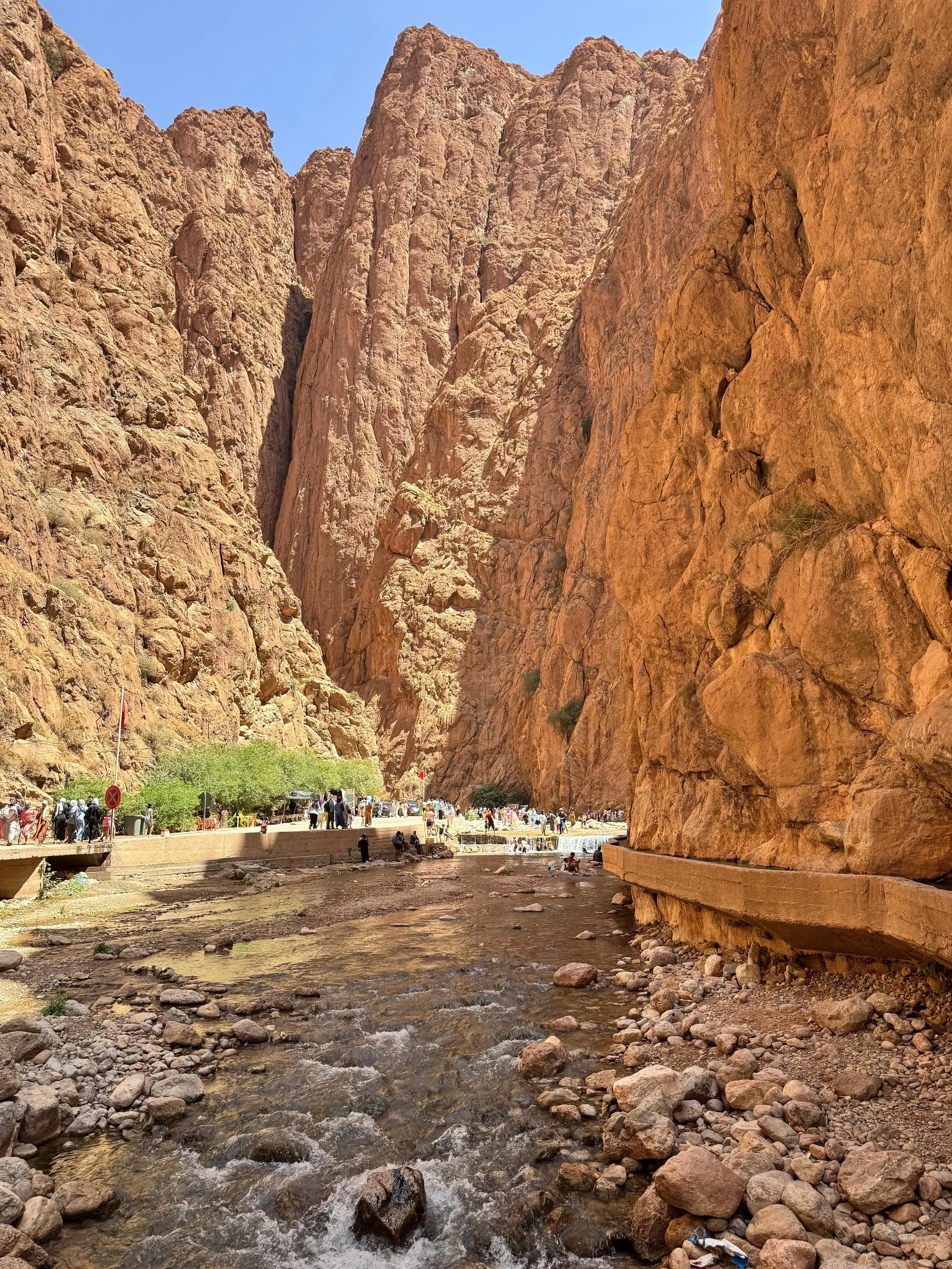 A narrow canyon with towering reddish-orange rock walls on either side, a small stream running through the rocky ground, and a pathway with tourists walking and relaxing, with a clear blue sky overhead.