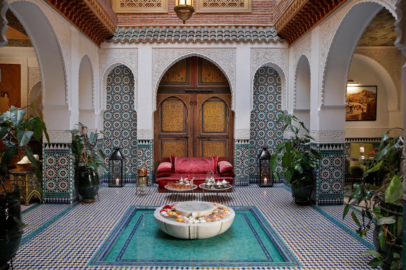 Interior of a Moroccan style lounge with a red sofa, a turquoise tiled floor, and ornate white walls with archways and intricate mosaic designs. A central fountain is decorated with flower petals, and there are large potted plants on either side.