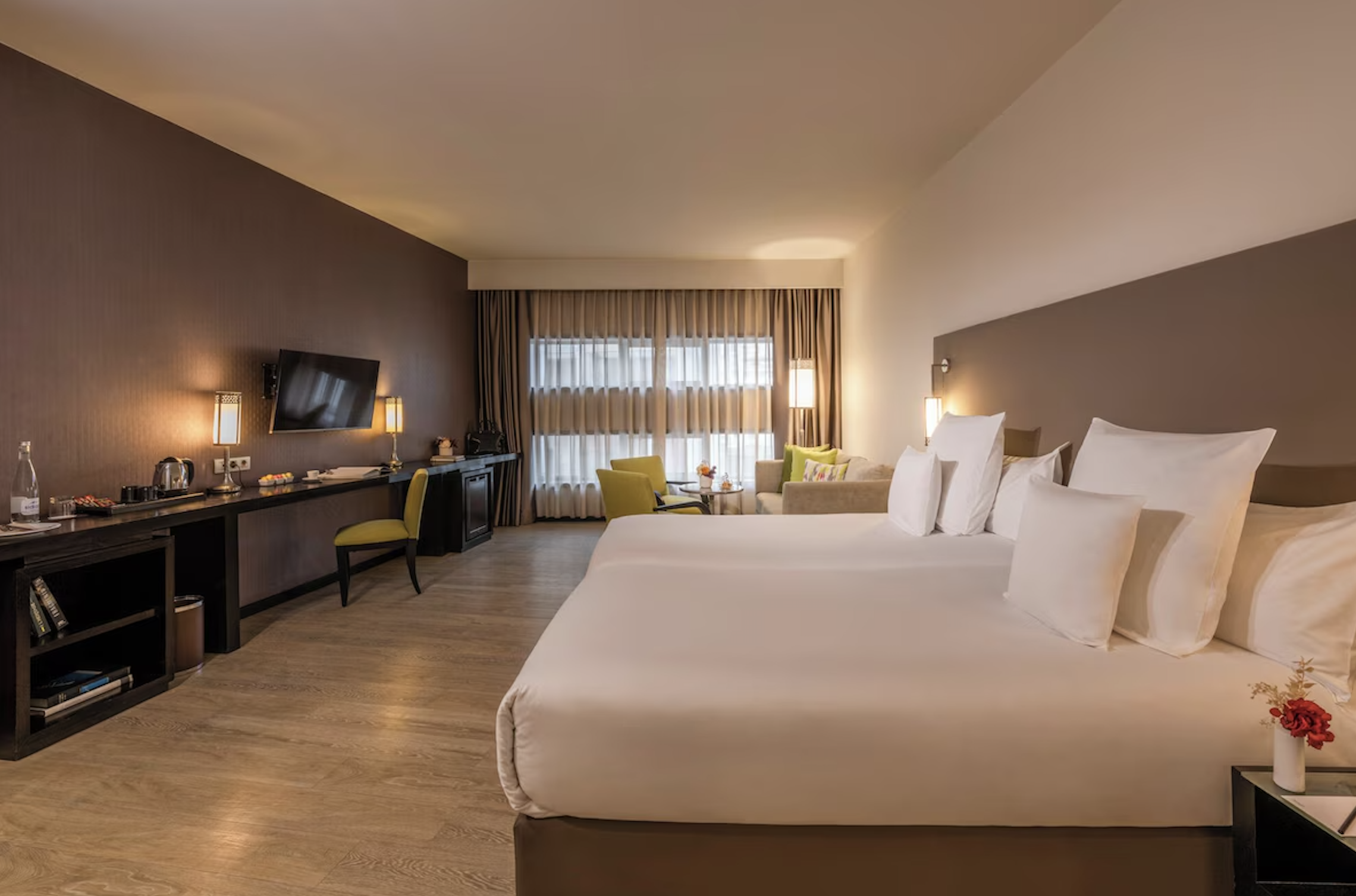 Spacious modern hotel room with two beds, neutral tones, desk and TV, seating area by a large window, and soft warm lighting creating a comfortable, contemporary feel.