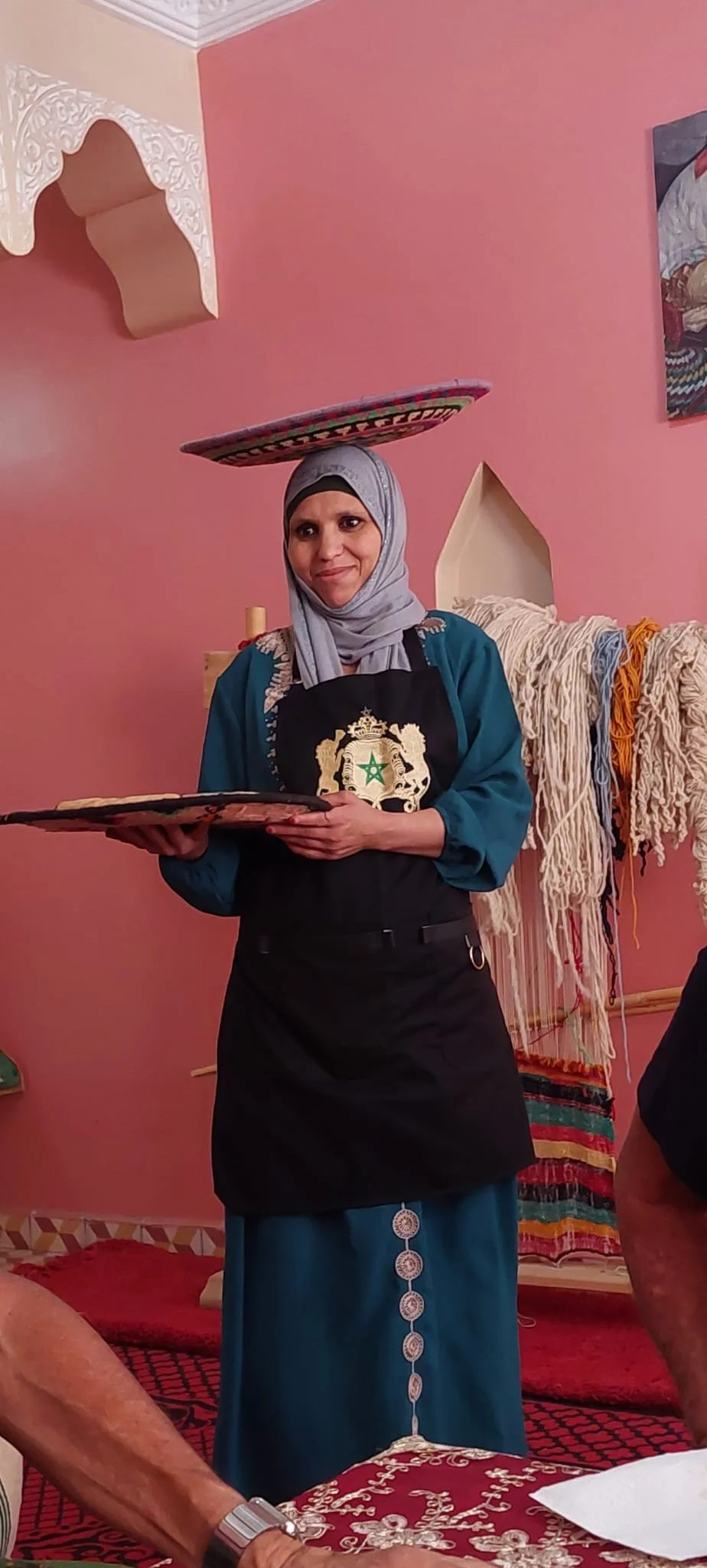 A woman wearing a hijab, black apron with a Moroccan emblem, and traditional dress balancing a decorated colored plate on her head and holding another plate in her hands, with colorful textiles hanging in the background.