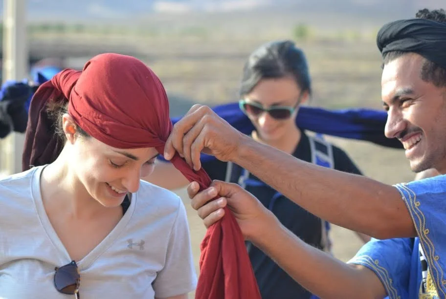A man is tying a red scarf around a woman’s head while smiling, with two other women smiling and observing in the background, outdoors.