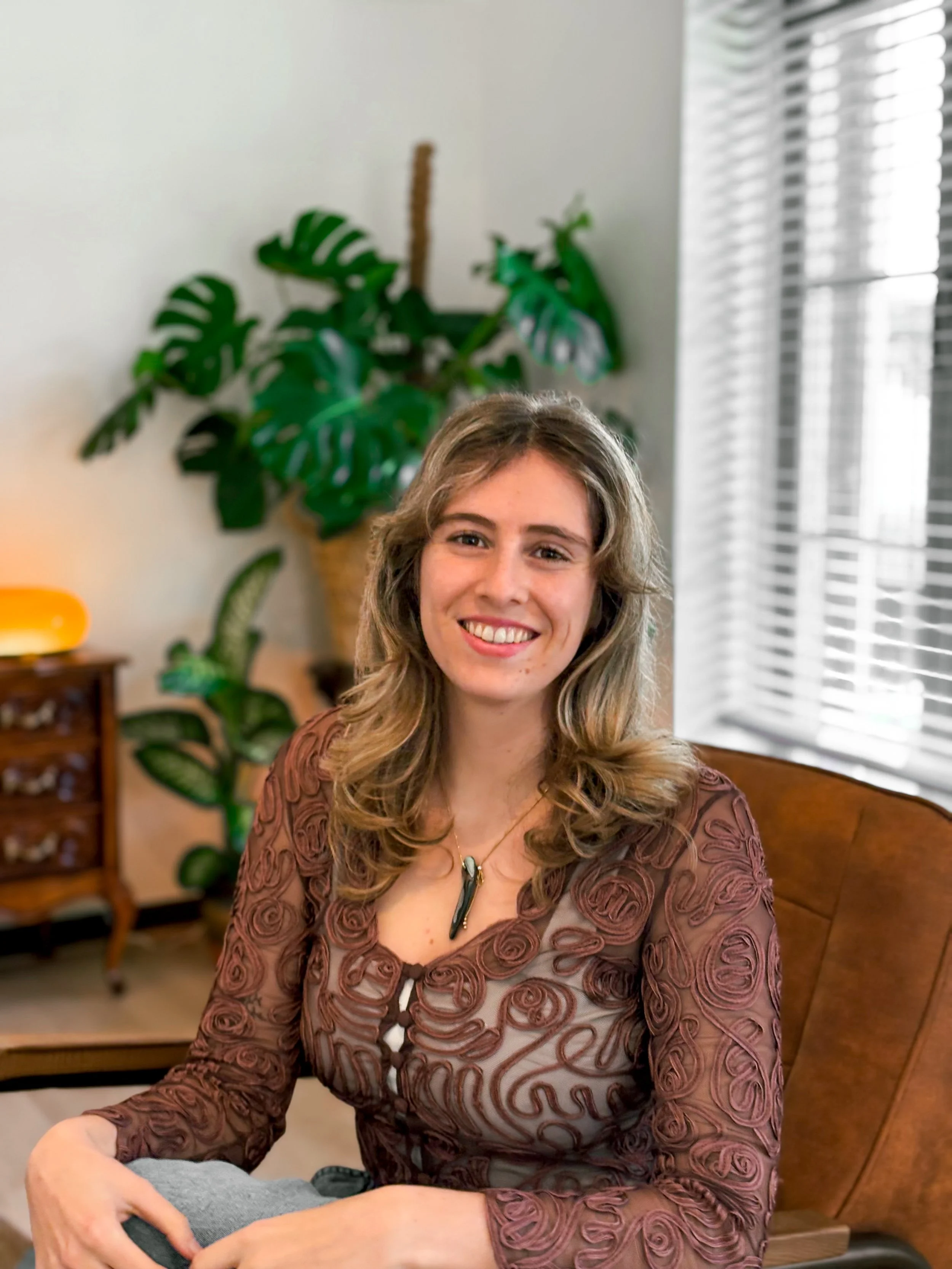 Een glimlachende vrouw met lang, golvend blond haar zit aan een marmeren tafel in een roze-witte koffiezaak, met bloemen in een glazen pot op tafel.