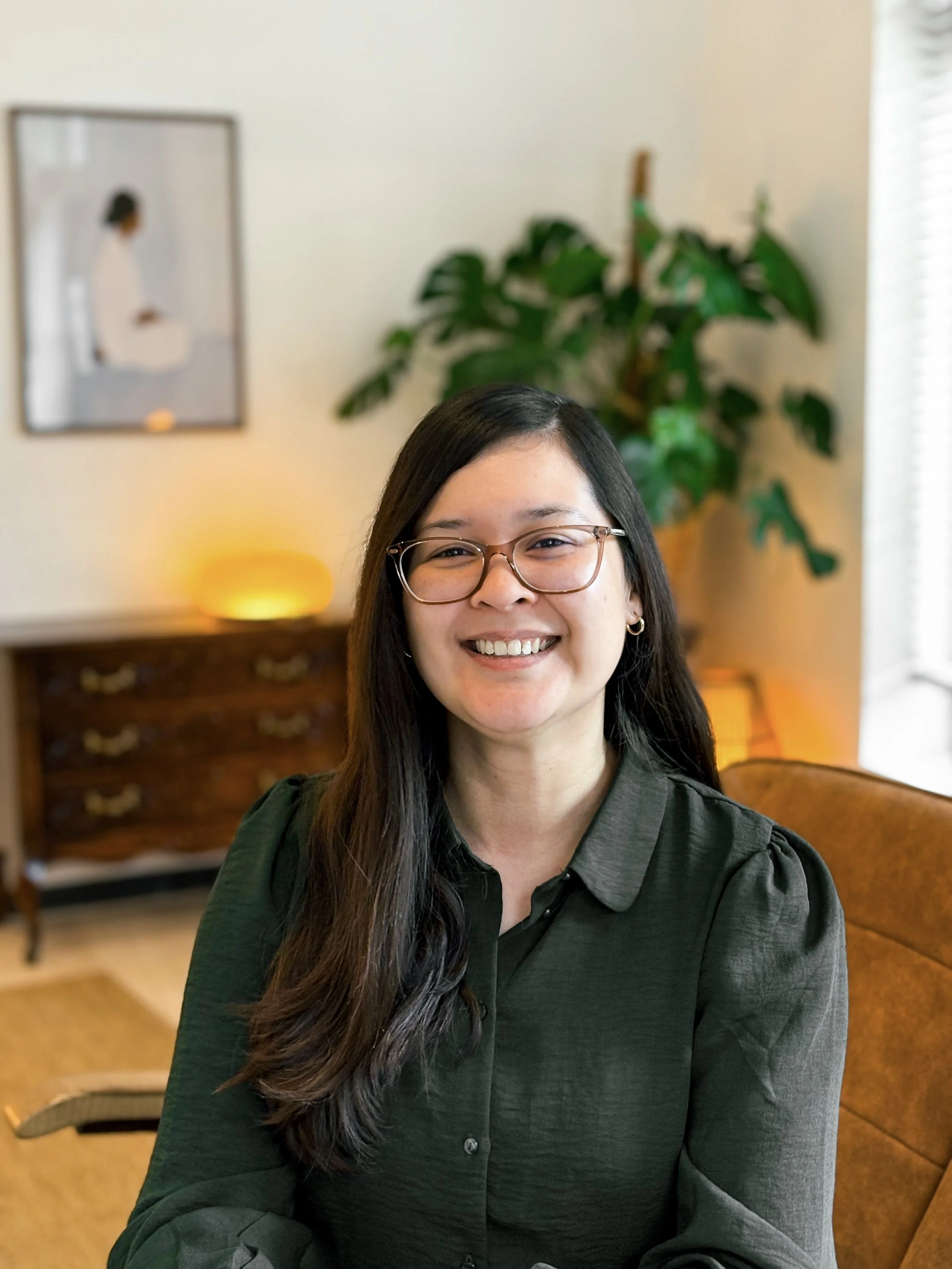 Een glimlachende vrouw met lang zwart haar, bril en een donkergroene blouse zit in een kantoor met planten en een schilderij op de muur.