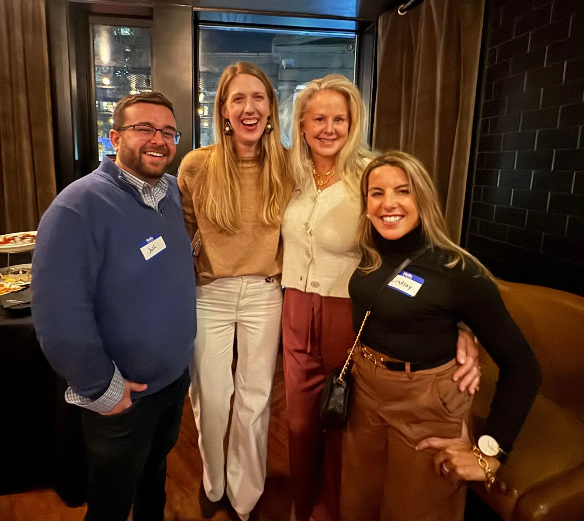 Four people smiling and posing together indoors, wearing name tags. They are standing close in a cozy, warmly lit room with a bar or food table visible in the background.
