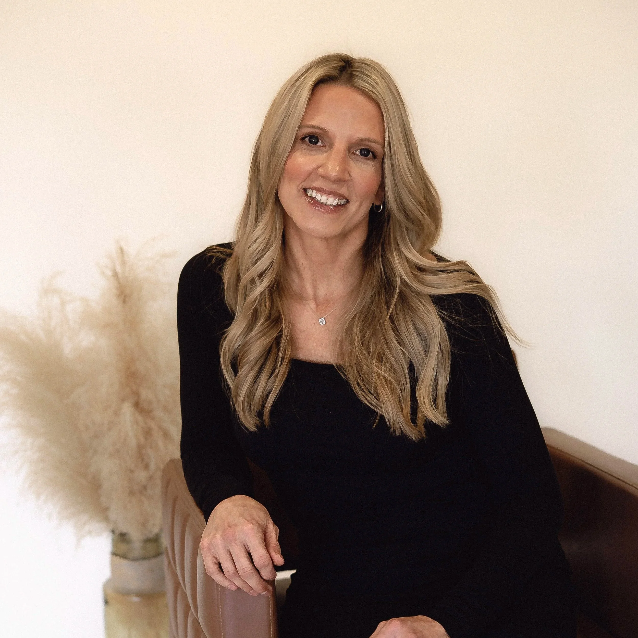 A smiling woman with long blonde hair wearing a black top, seated next to a beige chair, with a neutral background and pampas grass in a vase behind her.