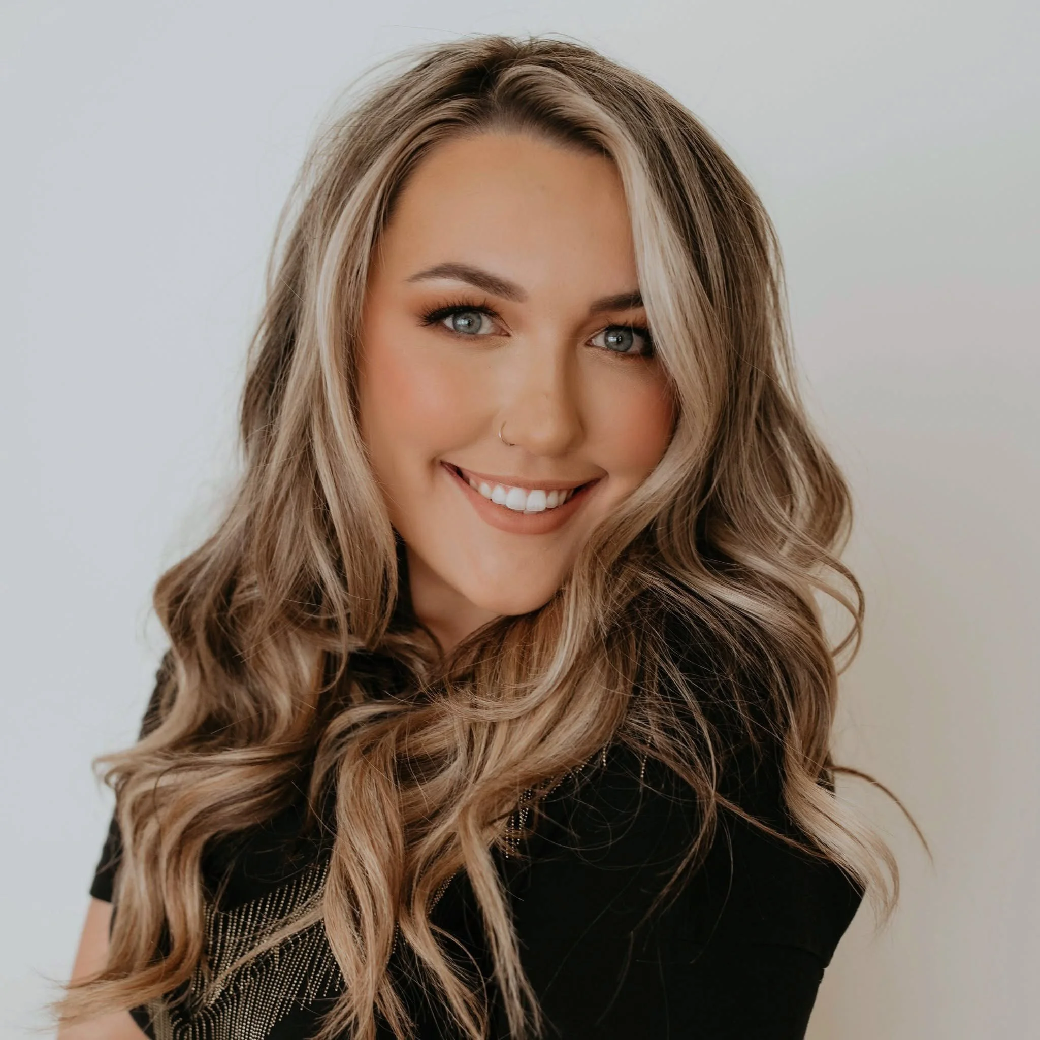 Close-up of a smiling woman with long wavy blonde hair, blue eyes, and a nose piercing, wearing a black top, against a plain light background.